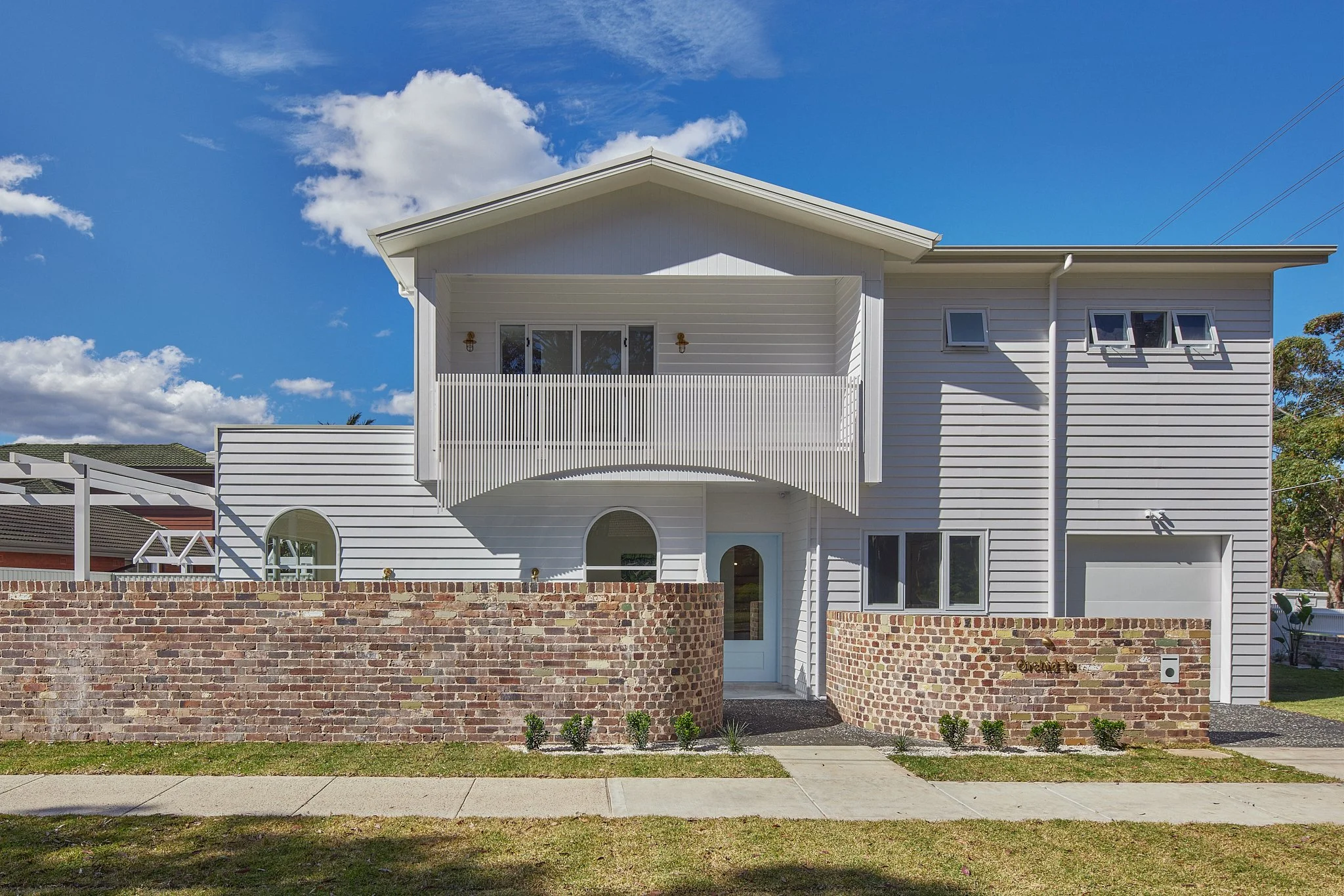 Front view of a modern white two-story house with a brick wall fence, small front yard, and sidewalk under a blue sky with scattered clouds.