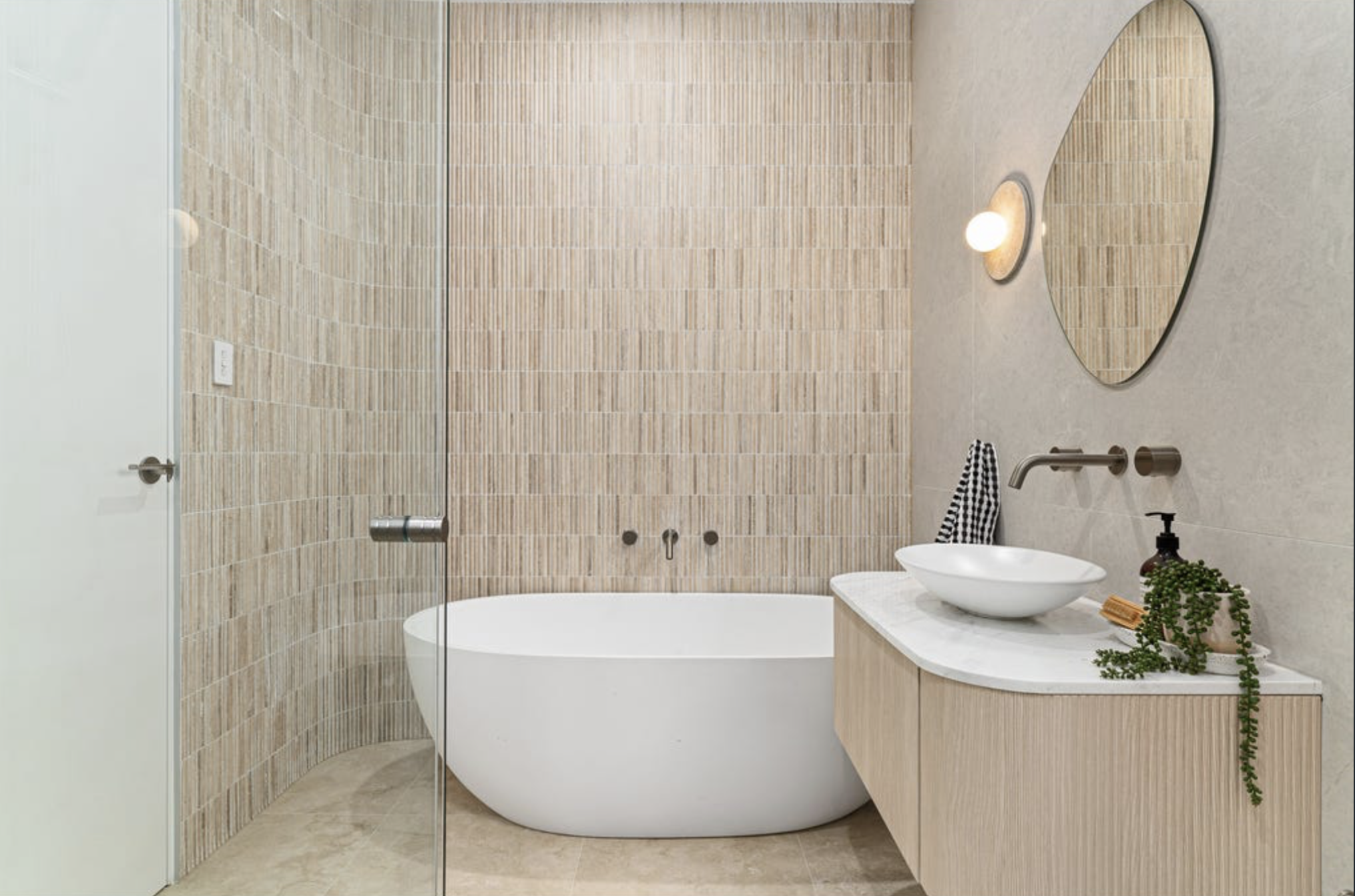 A modern bathroom featuring a white oval bathtub, a floating wooden vanity with a white vessel sink, a round mirror, and a wall-mounted light fixture. The décor includes a black-and-white striped towel and green plants, with beige tiled walls and floor.
