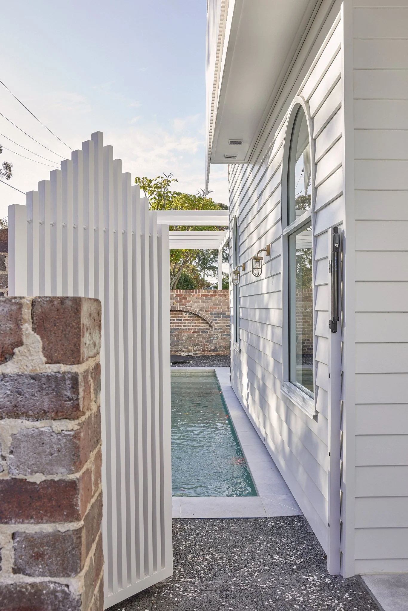 A narrow backyard with a small rectangular pond or pool beside a white house with horizontal siding, arched window, and various wall-mounted light fixtures. There is a white fence and brick wall in the background, with trees and a clear sky above.