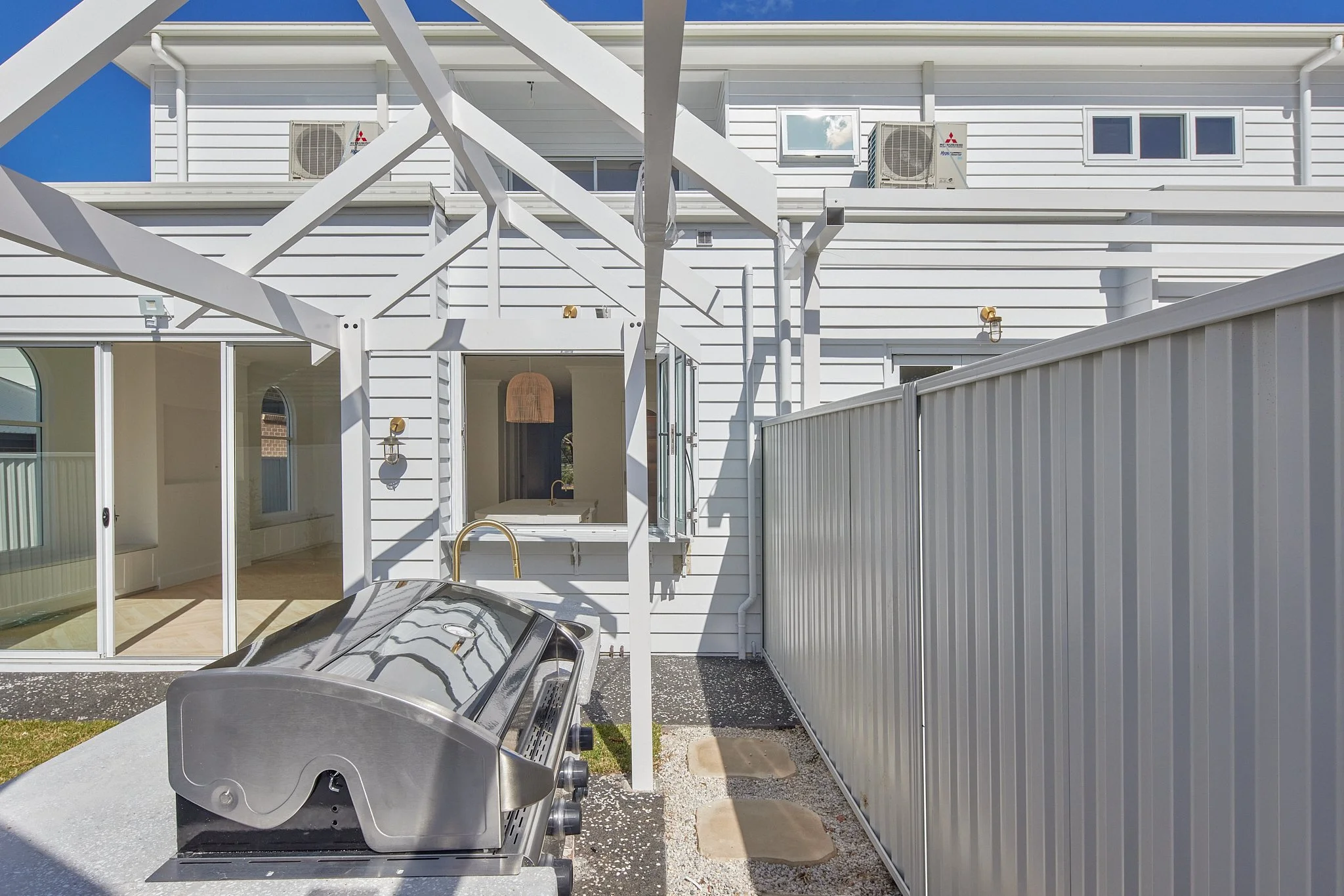 Backyard outdoor area with stainless steel grill, white fencing, sliding glass door, and a white house with air conditioning units on the upper level.