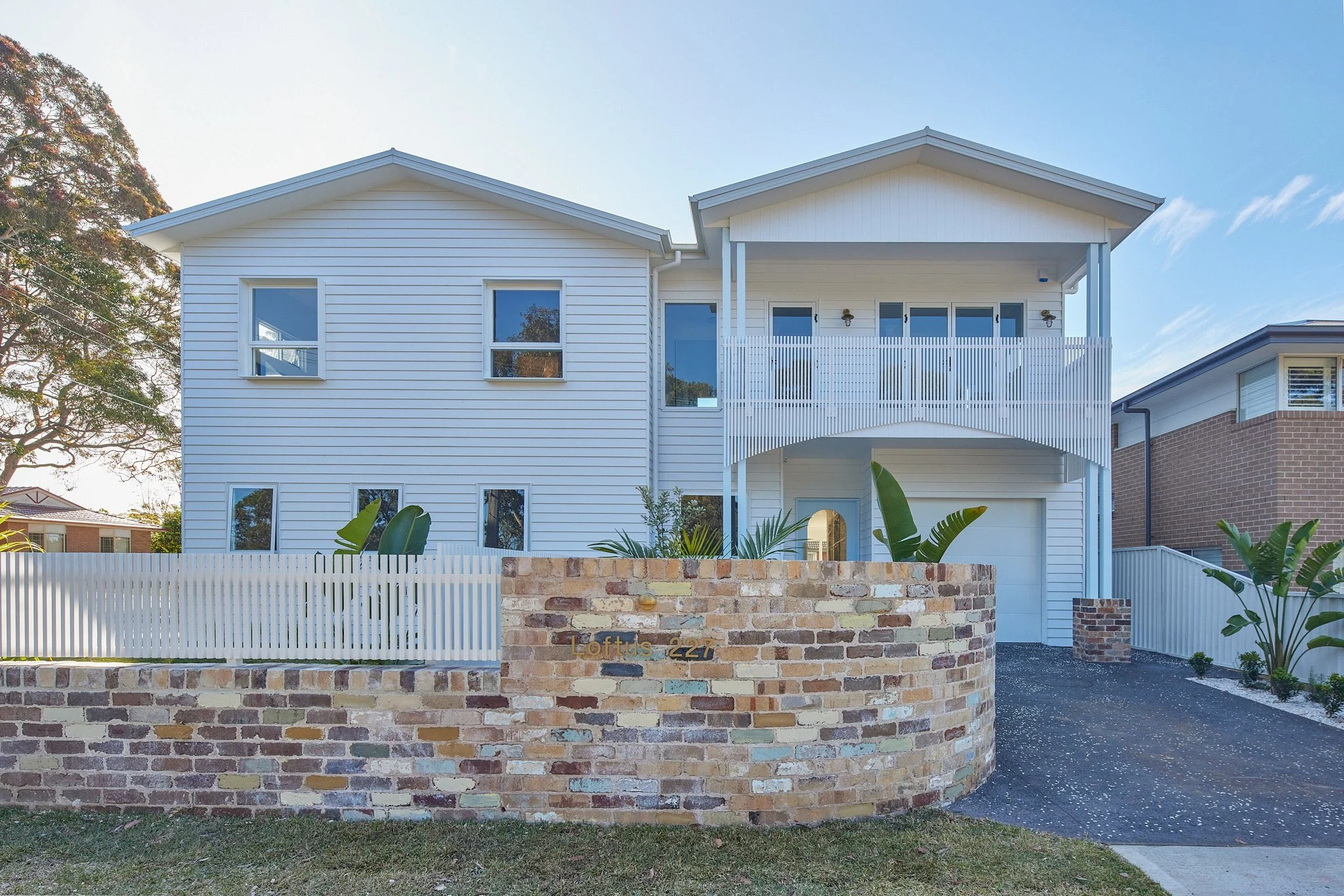 Front view of a modern two-story white house with a brick and white picket fence, lush greenery, and a driveway.