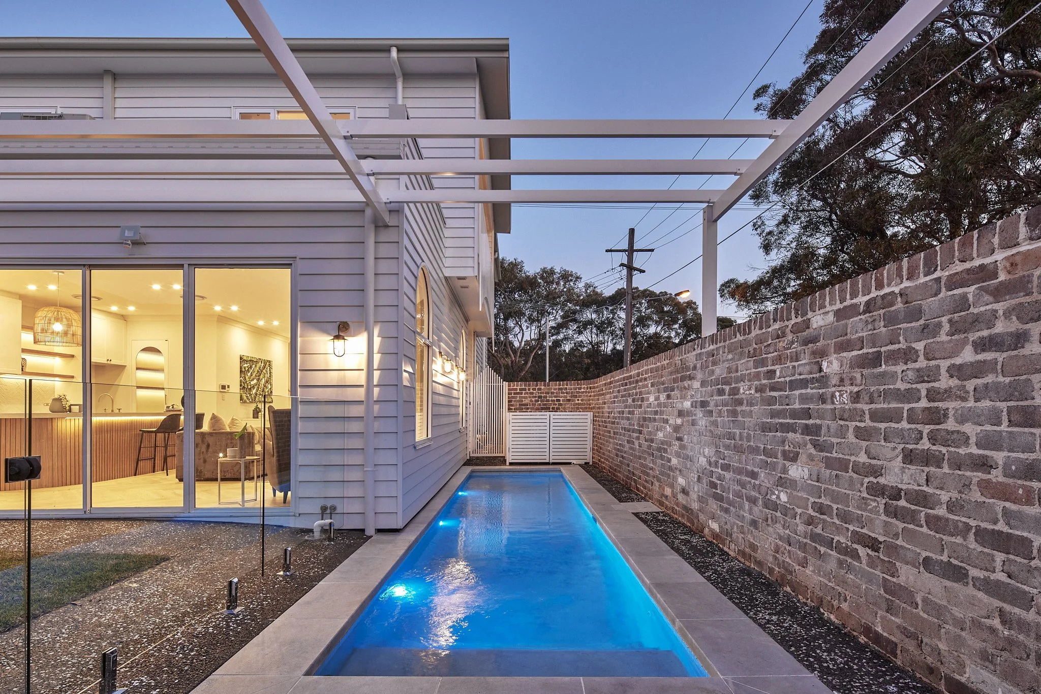 A backyard with a narrow illuminated swimming pool, a brick wall on one side, and a white house with large glass sliding doors providing a view of the interior living space, at dusk.