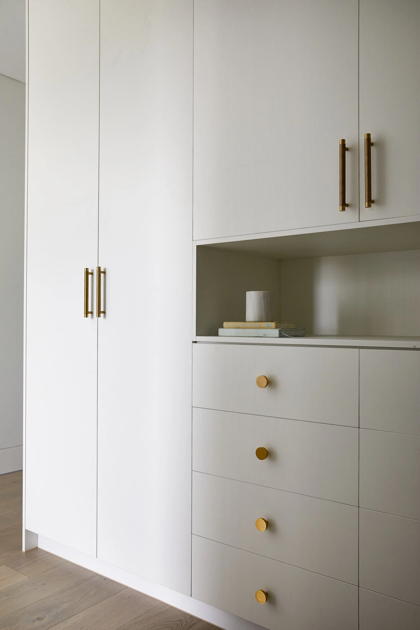 Close-up of a white built-in cabinet with gold handles and knobs, featuring four drawers with round knobs, a small open shelf with a stack of books and a white sculpture, and tall cabinet doors.