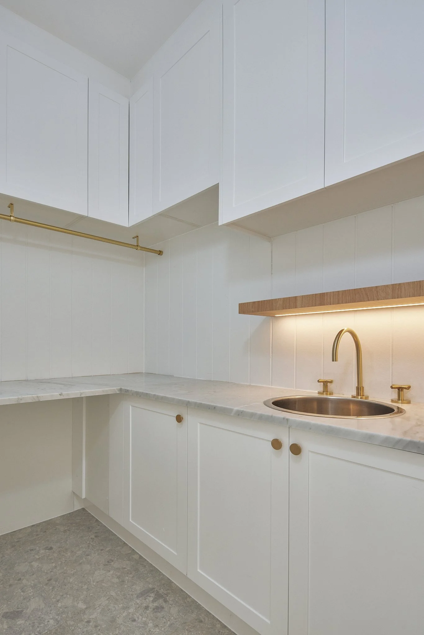 Minimalist white kitchen with white cabinetry, marble countertop, gold fixtures, and wooden shelf with underlighting.