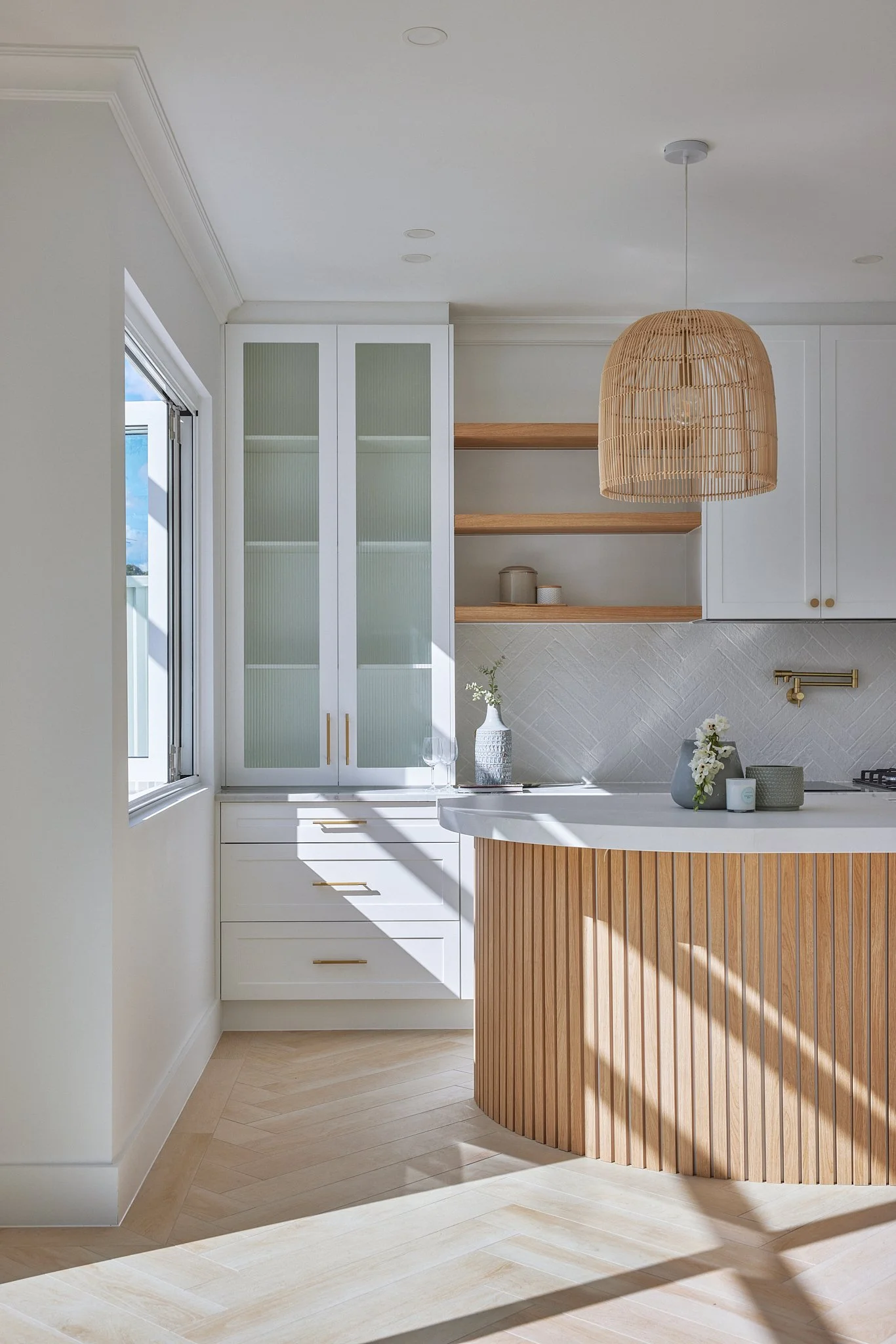 Modern kitchen with white cabinets, open wooden shelves, and a curved wooden island with a white countertop. A wicker pendant light hangs from the ceiling, and there is natural light coming through a window on the left.