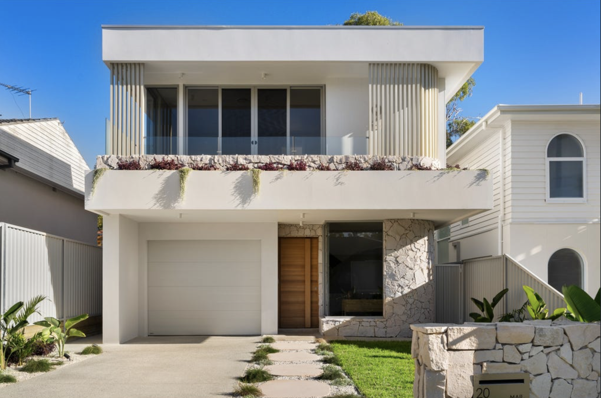 Modern two-story house with white exterior, stone accents, and a garage door on the left. A wooden front door and large front window are visible, with a small lawn and stepping stones leading to the entrance. The balcony on the upper level has a glass railing and balcony box with plants.