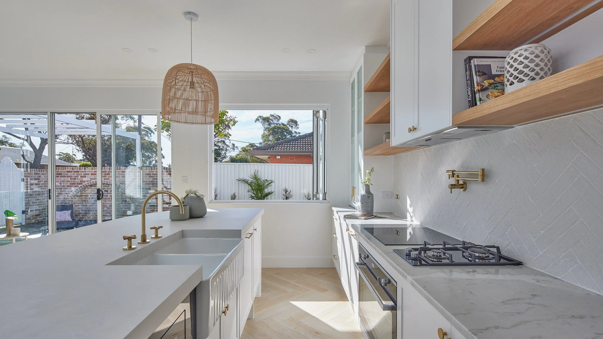 Bright modern kitchen with white cabinets, a marble countertop with a sink, gas stove, open wooden shelves, and large window showing backyard with trees and brick wall.