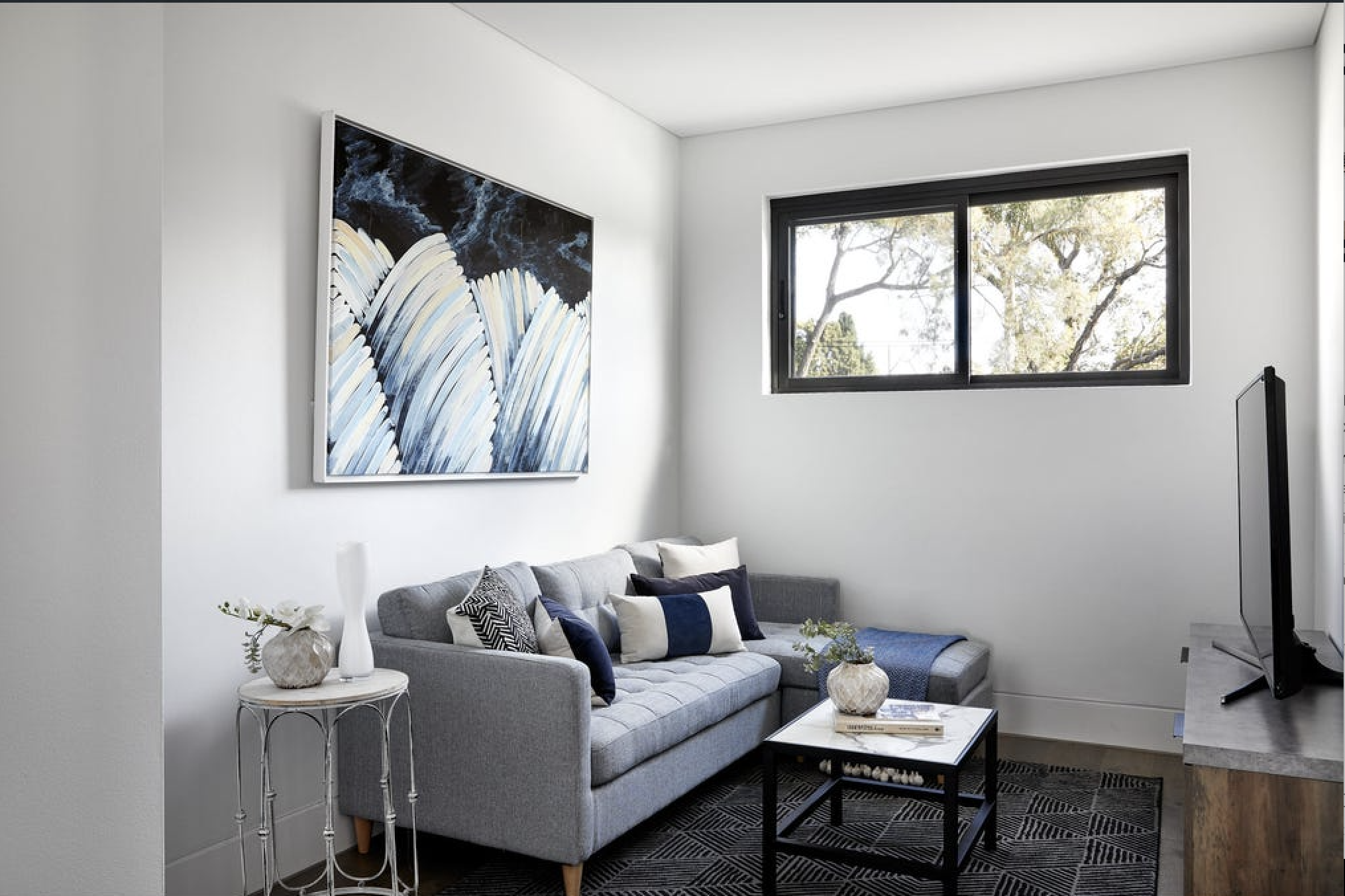 Modern living room with gray sofa, black and white pillows, a side table with decorative vases, a coffee table with a vase and books, a large abstract painting, a flat-screen TV, and a window showing trees outside.