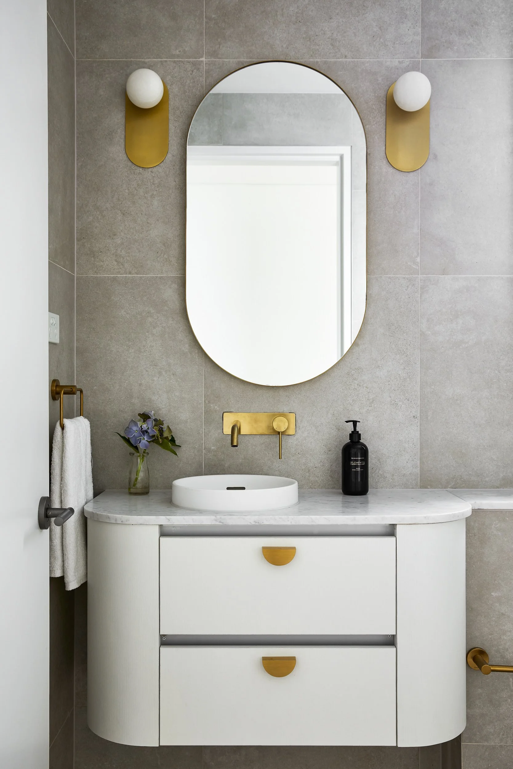 Modern bathroom vanity with a white marble countertop, oval vessel sink, and a gold faucet. A large oval mirror is mounted on a gray tiled wall, with two white globe wall sconces. A small vase with purple flowers and black soap dispenser are on the c