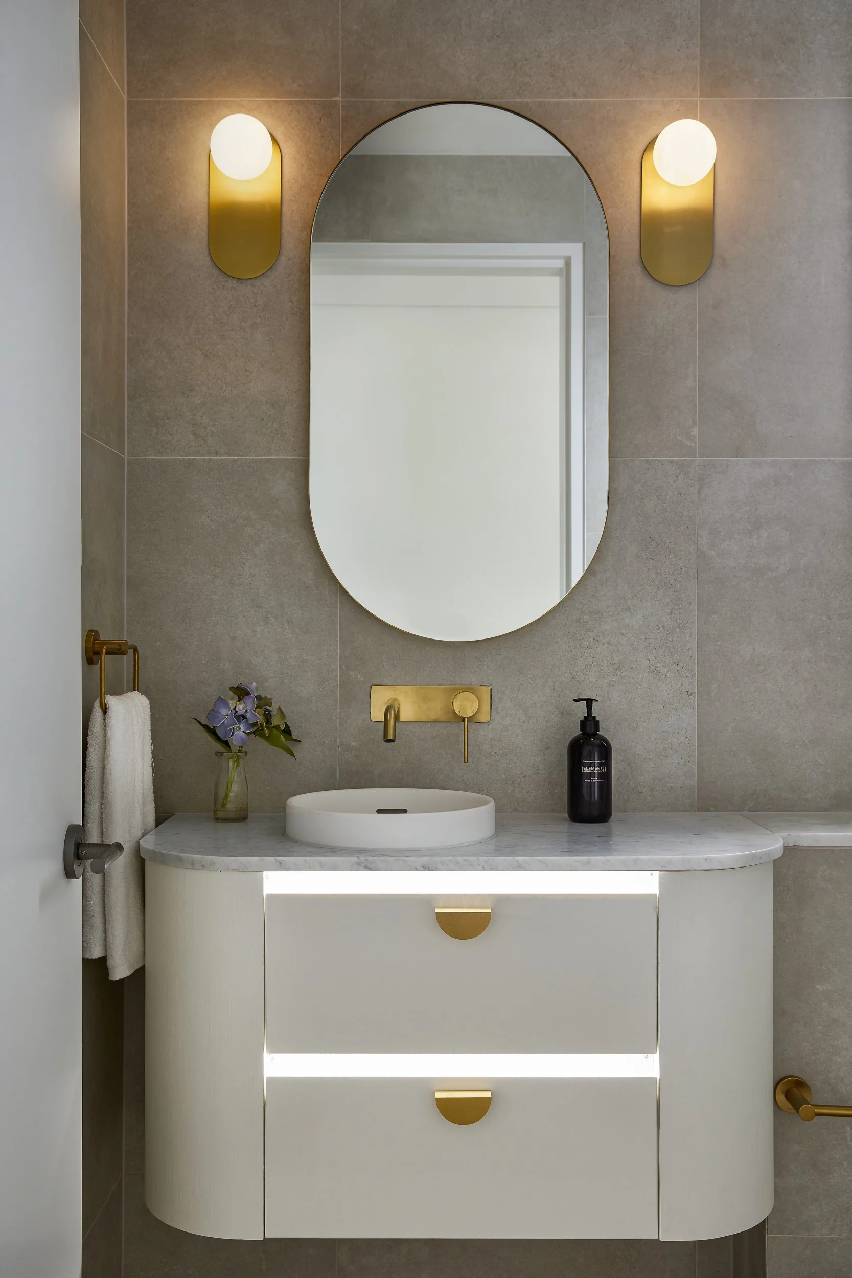 Modern bathroom vanity with a white curved cabinet, a marble countertop, and a vessel sink. A large oval mirror is mounted above with two gold wall sconces on either side. A black soap dispenser and a small vase with purple flowers are on the counter