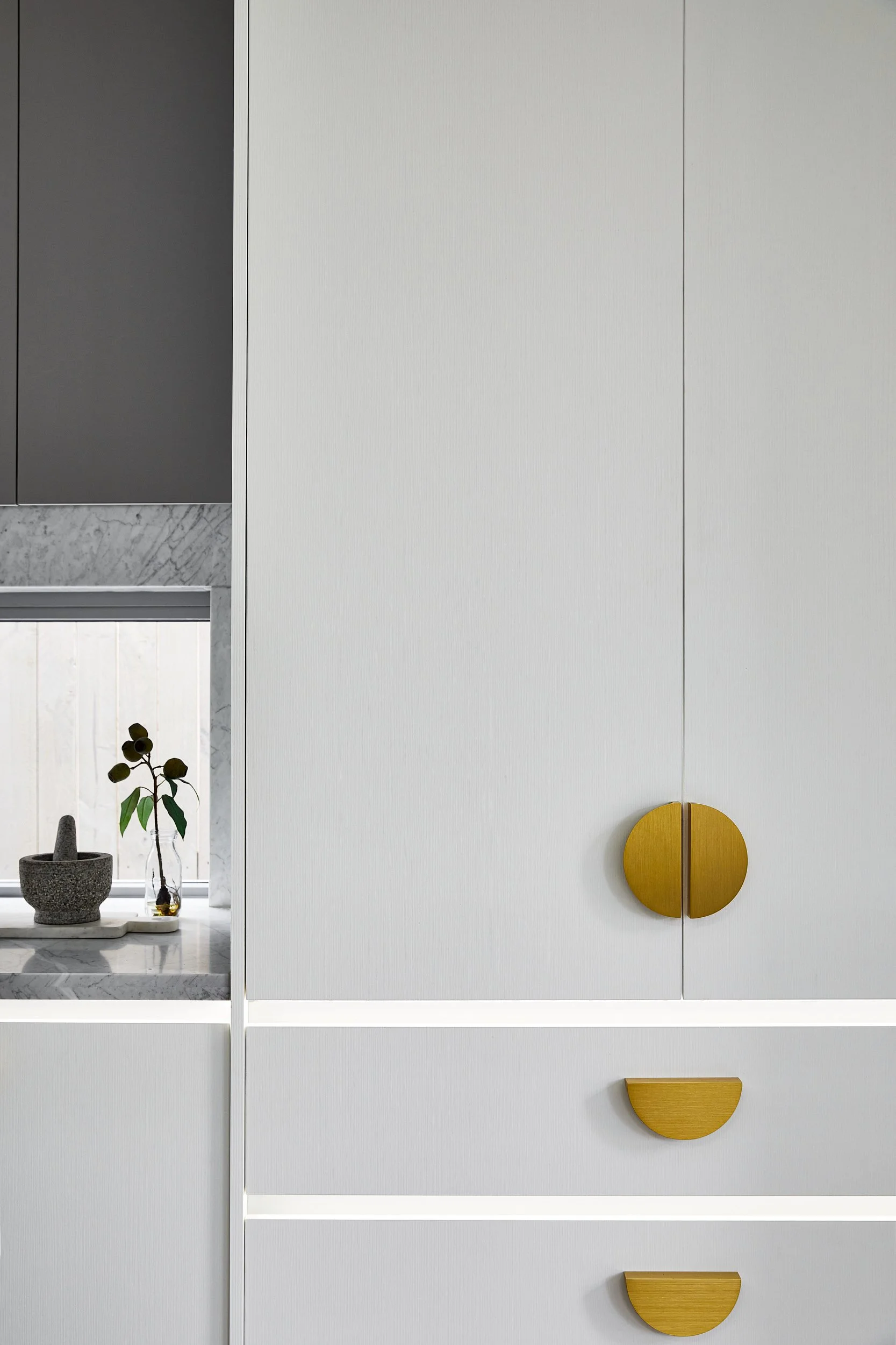 Close-up of a modern white cabinet with round, half-moon gold handles and a small marble shelf holding a potted plant and a mortar and pestle.