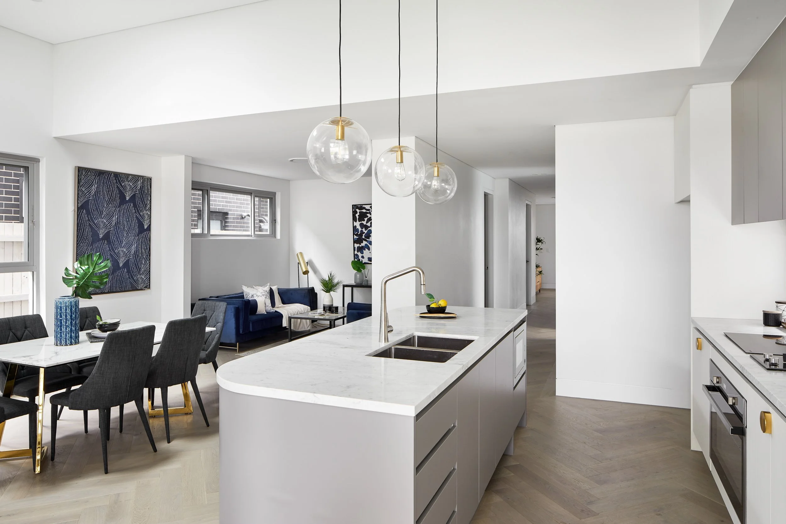 Modern open-concept kitchen and living room with white walls, marble countertops, and dark furniture, decorated with blue and green accents, and pendant lights hanging over the kitchen island.