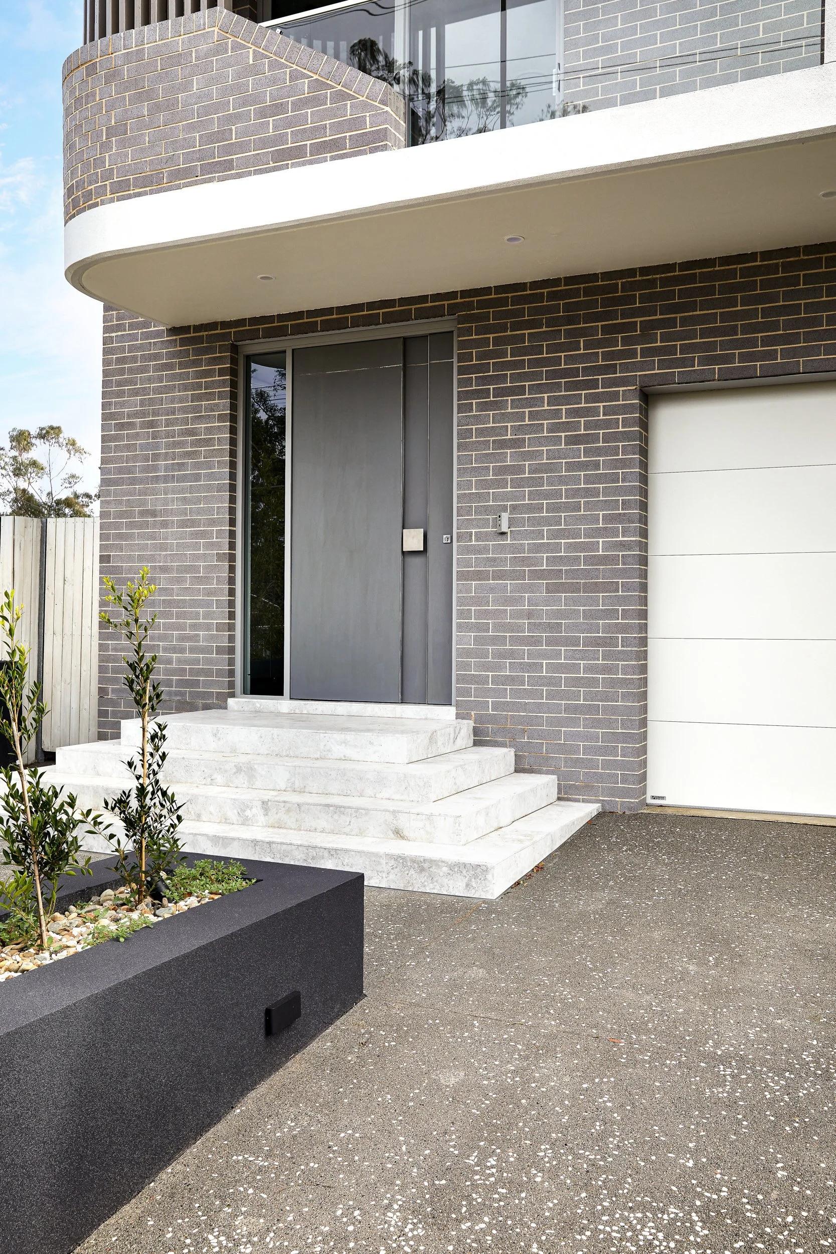 Modern house entrance with dark metal door, white concrete steps, gray brick exterior, white garage door, and a planter with small green plants and rocks.