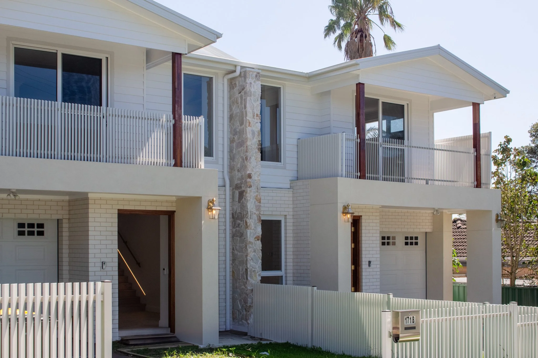 Modern multi-story house with white siding, stone chimney, and balconies with vertical metal railings, surrounded by a white picket fence and palm trees in sunny weather.