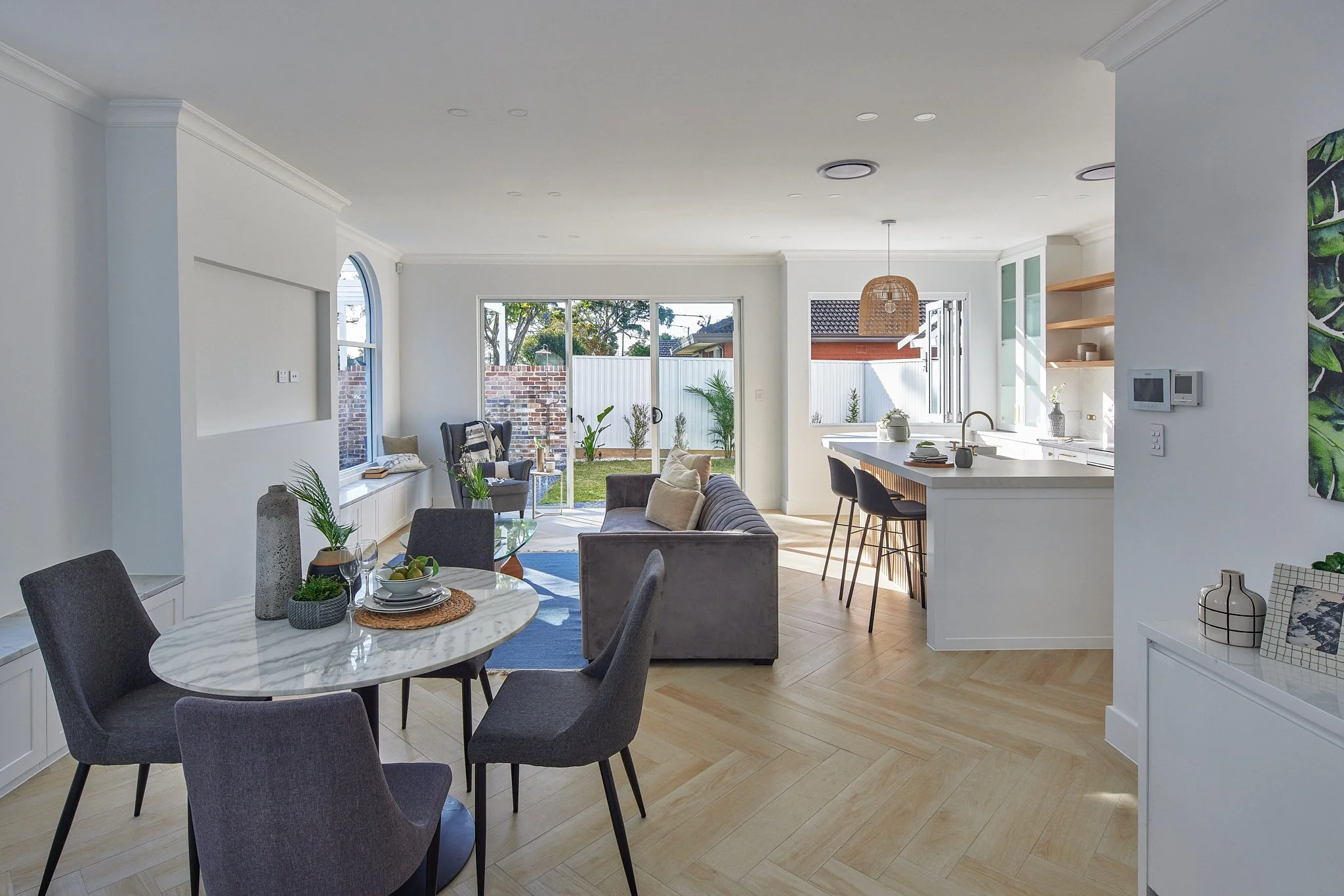 Open-concept living and dining area with modern decor, light wood flooring, grey sofa, round marble dining table with six grey chairs, bright kitchen with white cabinets, barstools, and large windows providing natural light.