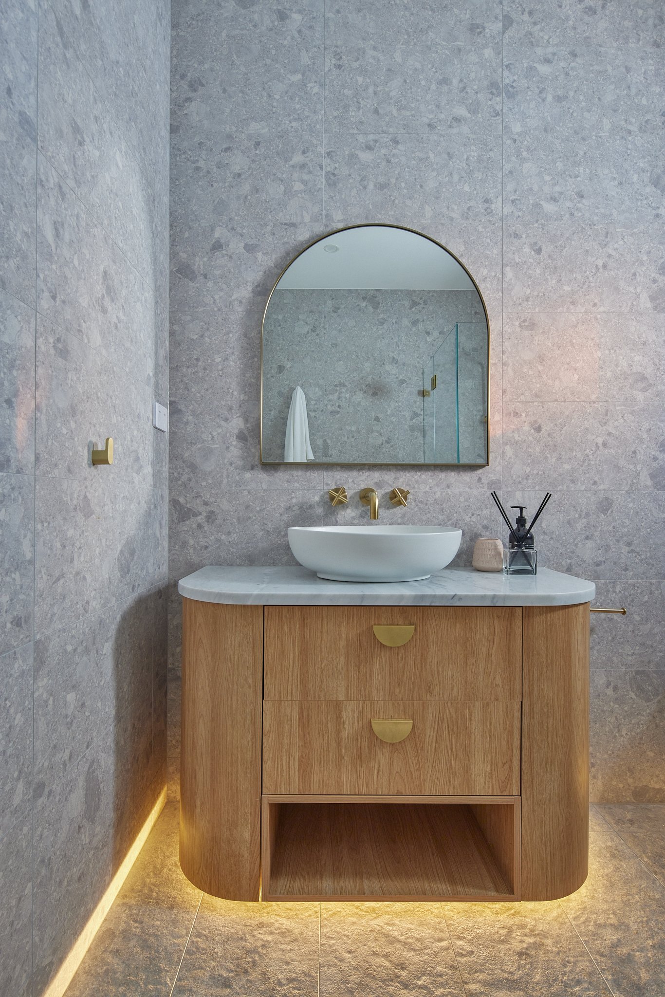Modern bathroom vanity with a white vessel sink, gold fixtures, a marble countertop, a wood cabinet, and an arched mirror on a gray stone wall, illuminated by warm lighting at the base.