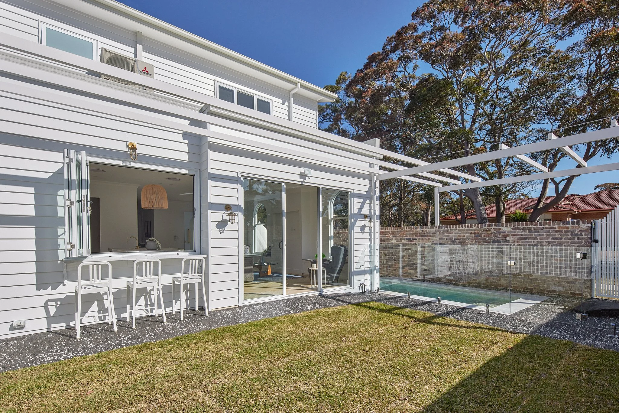 Backyard of a white house with a glass door, outdoor seating, and a small pool with glass fencing, surrounded by a grass lawn and trees.