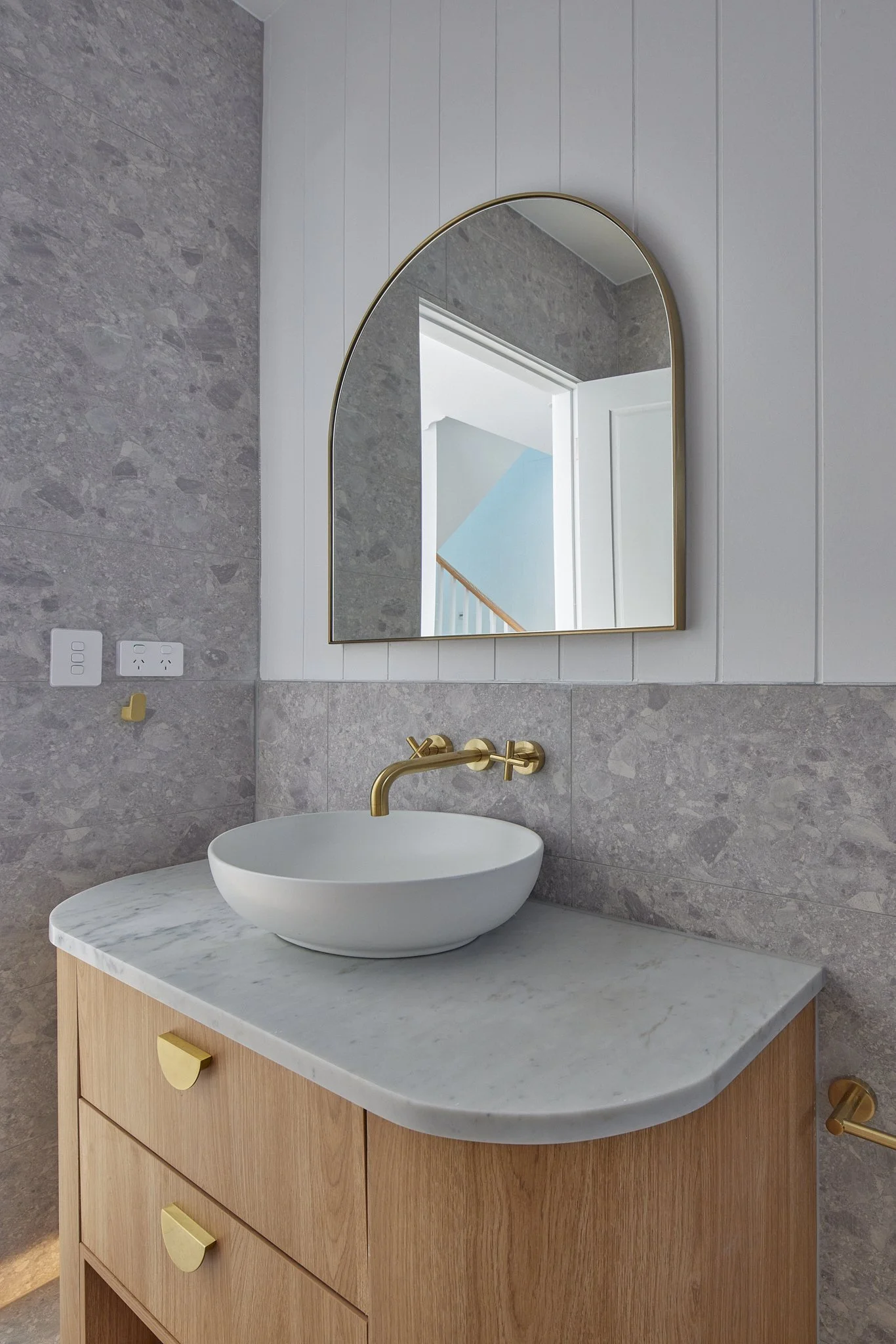A bathroom vanity with a white vessel sink, gold faucet, and a wooden cabinet topped with a marble countertop. Above is an arched mirror reflecting a doorway and staircase.