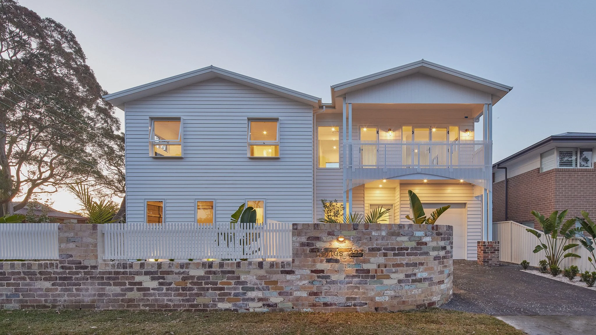 Three-story white house with a brick and white fence, a driveway, and lush greenery in front, illuminated at dusk.