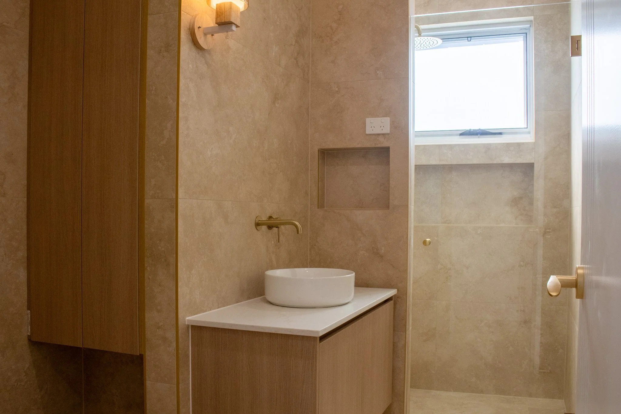 A modern bathroom with beige tiled walls and floor, a white countertop with a round vessel sink, a wall-mounted faucet, a wooden cabinet, a small wall-mounted light fixture, a window, and a separate shower area with a built-in niche and a rainfall showerhead.