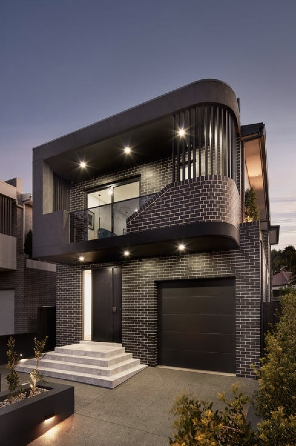 Modern two-story house with black brick exterior, illuminated outdoor lights, a garage, and stairs leading to a front door, during twilight.