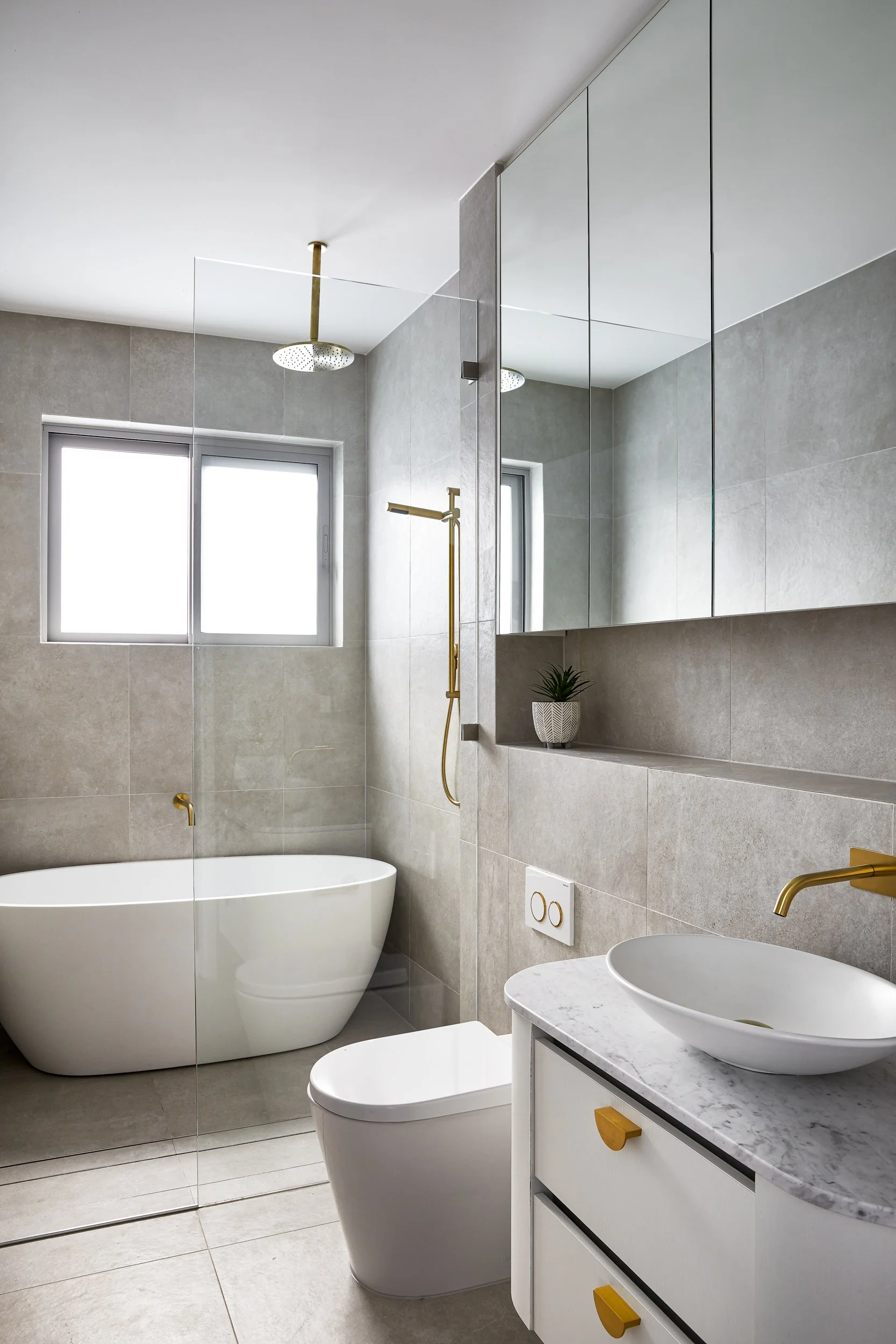 Modern bathroom with a freestanding bathtub, a wall-mounted toilet, a white vanity with a marble countertop and a vessel sink, gold fixtures, a window with frosted glass, a large mirror, and a small potted plant.