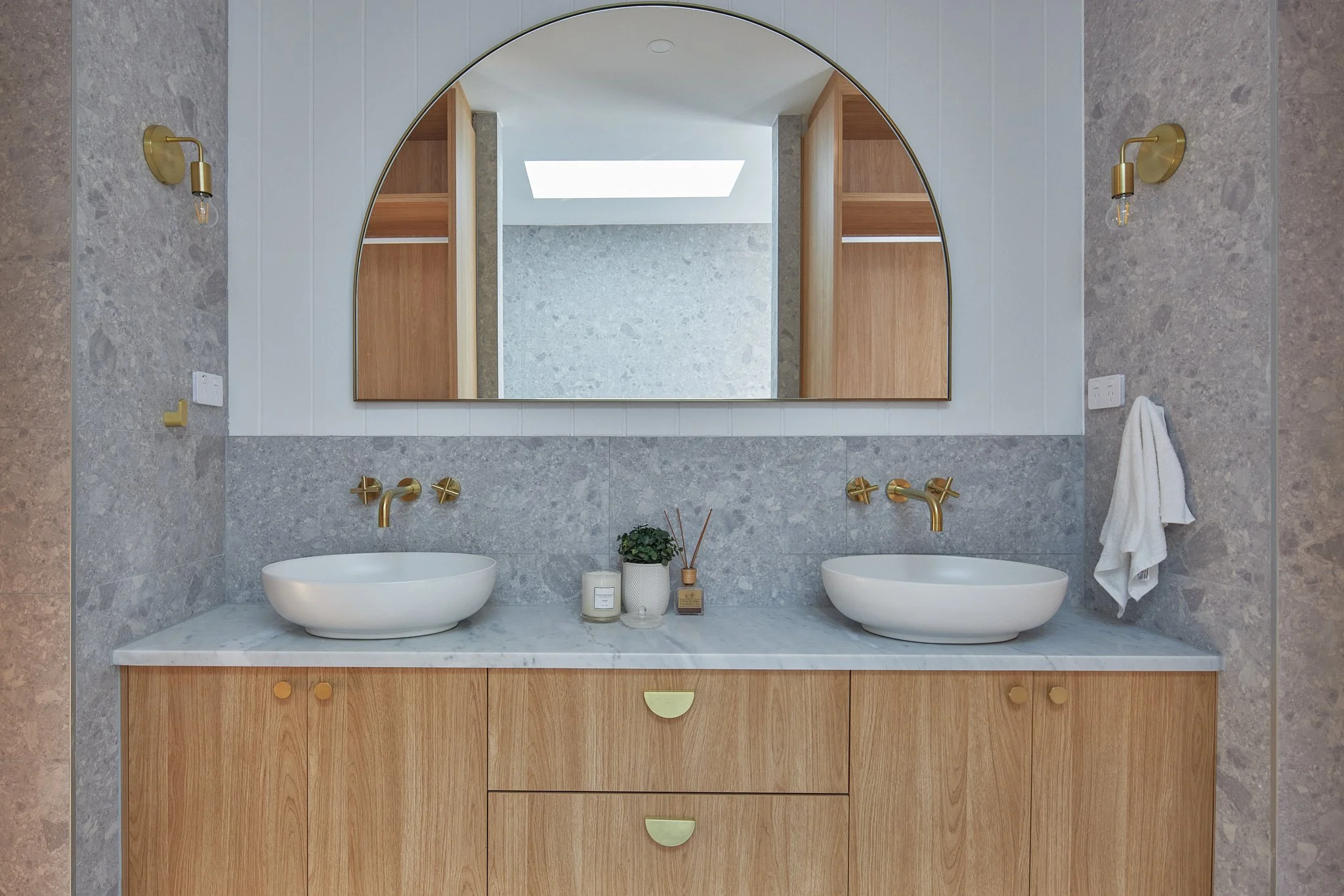 A modern bathroom vanity with a large mirror, two vessel sinks with gold faucets, a wooden cabinet with three drawers, and decorative items such as a plant, diffuser sticks, and candles.