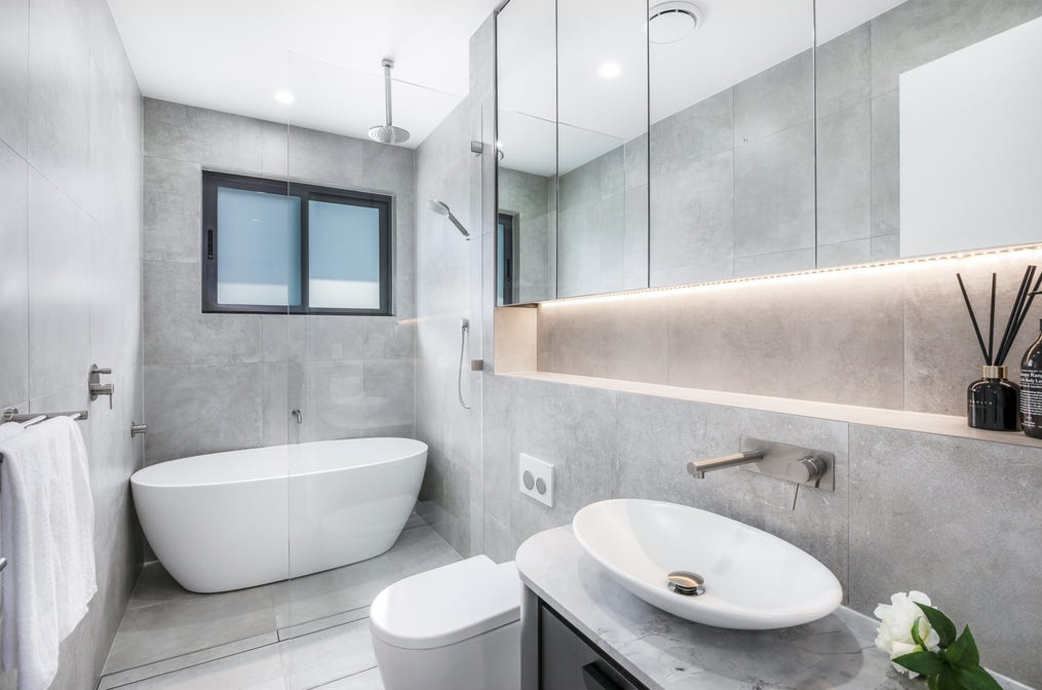 Modern bathroom with gray tiled walls and floor, a white freestanding bathtub, gray vanity with a vessel sink, large mirror with built-in lighting, window, and minimal decor including a black reed diffuser and white flowers.