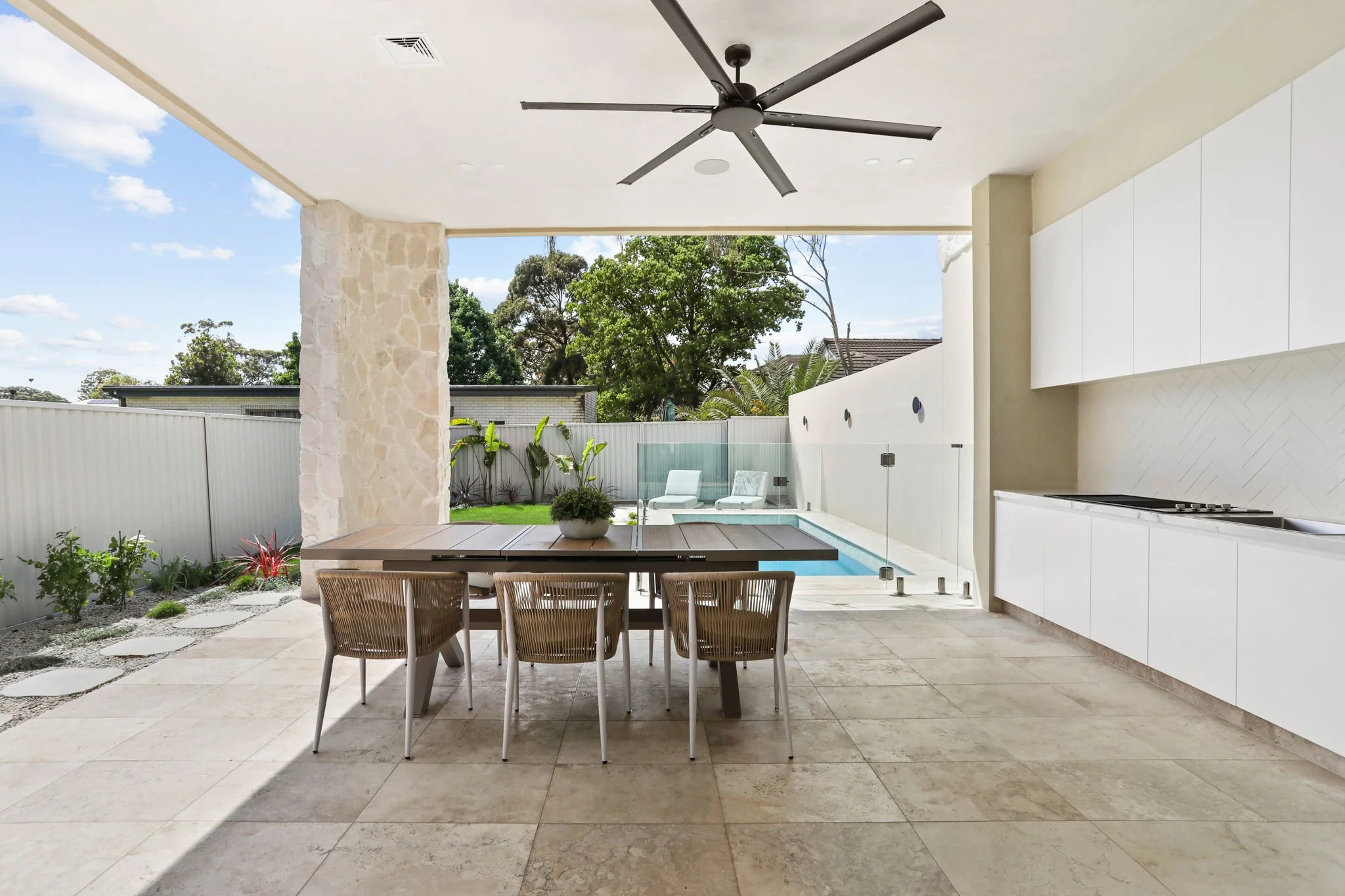 Covered outdoor patio with a dining table and six chairs. In the background, there's a small pool with a glass fence, lounge chairs, and green trees and plants. The area is bright and sunny.