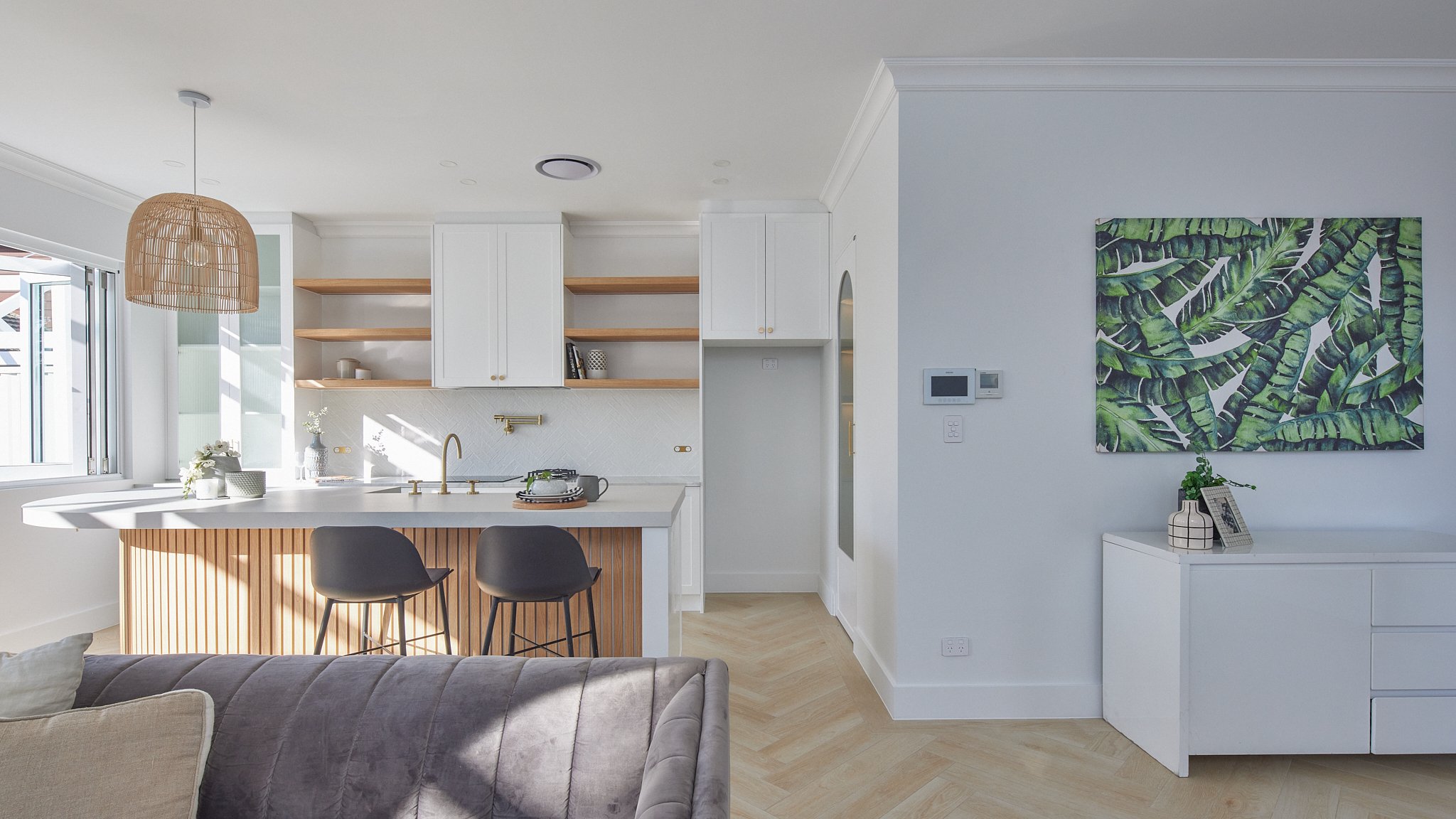 Modern kitchen with white cabinetry, open wooden shelves, and a white countertop island with black barstools. A rattan pendant light hangs from the ceiling, and there's a large window letting in natural light. A beige sofa with a light-colored pillow
