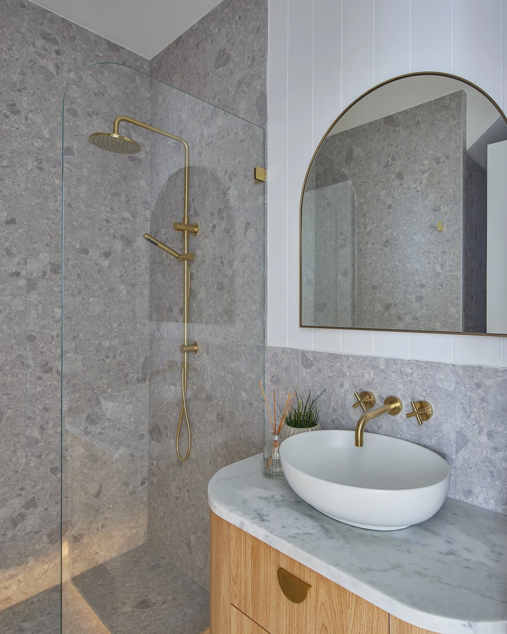 Modern bathroom with a walk-in shower, gold fixtures, a white vessel sink on a wood vanity, a round mirror, and a decorative plant