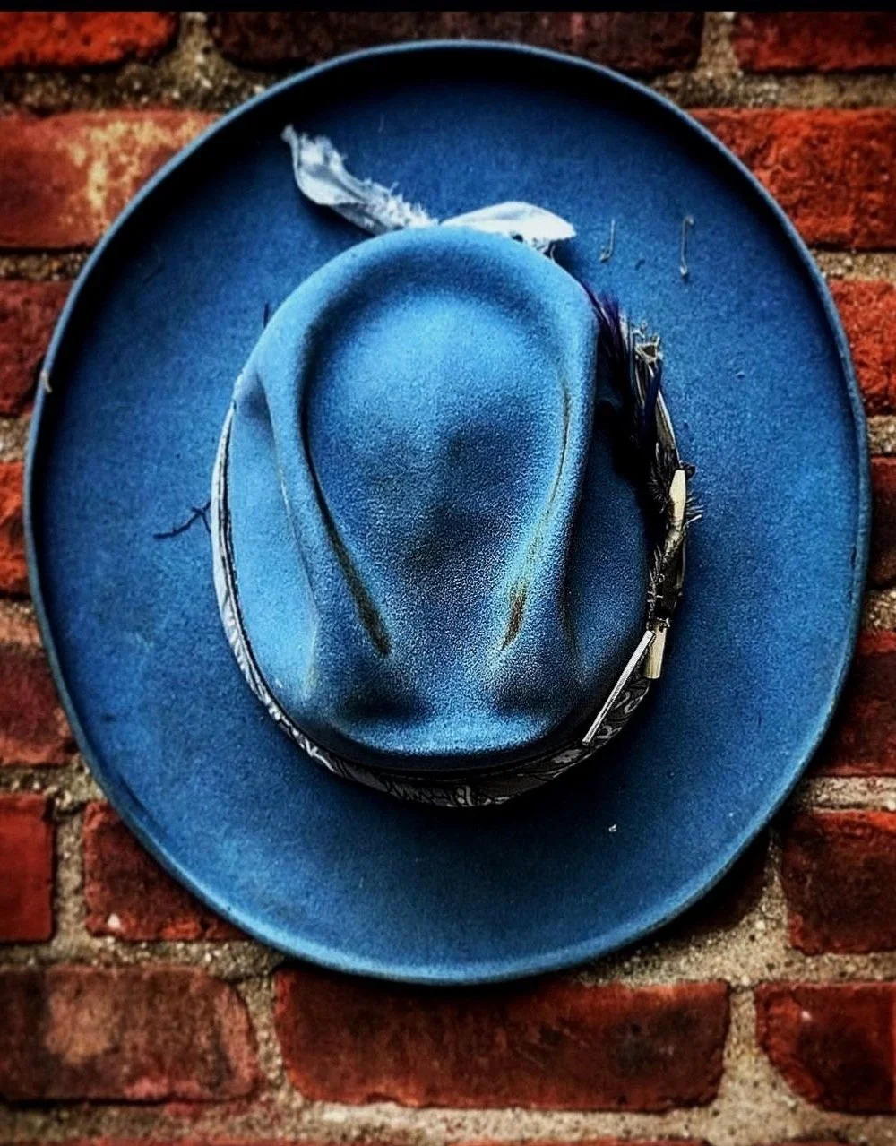 A blue fedora hat with a feather, resting on a matching blue hatband, placed on a blue hat. The hats are on a brick wall background.