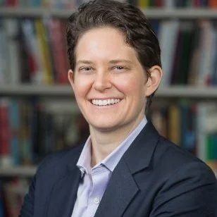 Portrait of a person with short, curly dark hair, wearing a navy blazer and a light blue shirt, smiling in front of a bookshelf filled with books.