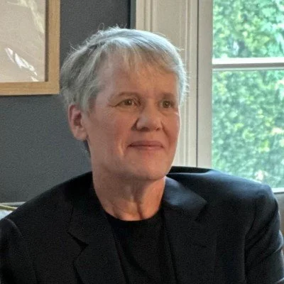 Close-up of an older woman with short gray hair, wearing a black blazer, sitting indoors near a window with green trees visible outside.