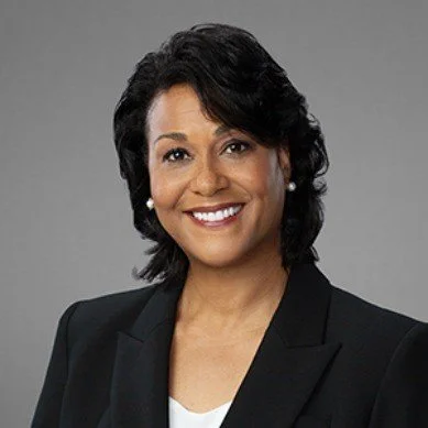 Professional woman with short black curly hair, wearing a black blazer and white top, smiling against a gray background.