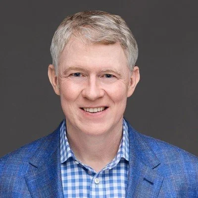 Headshot of a smiling middle-aged man with short, gray hair, wearing a blue checkered shirt and a blue blazer against a gray background.