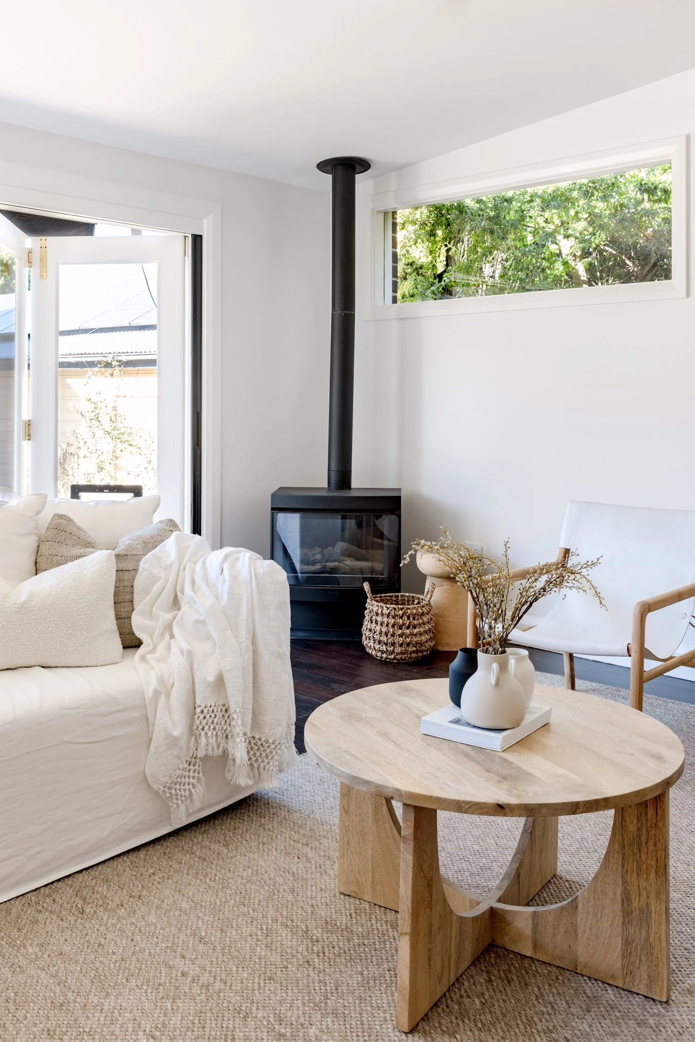 Living room with a white sofa, beige cushions, a wooden coffee table with vases and dried flowers, a white chair, a wood-burning stove, and large windows with greenery outside.