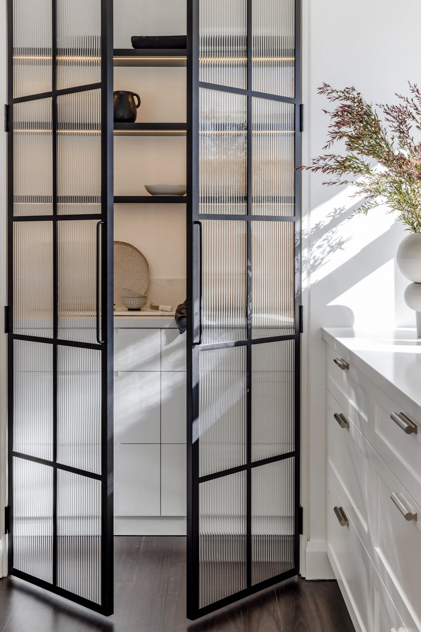 A black metal and glass cabinet with three shelves, partially open, situated next to a white kitchen counter with a potted plant on it. Items inside the cabinet include a black pitcher, a bowl, and a large tray.