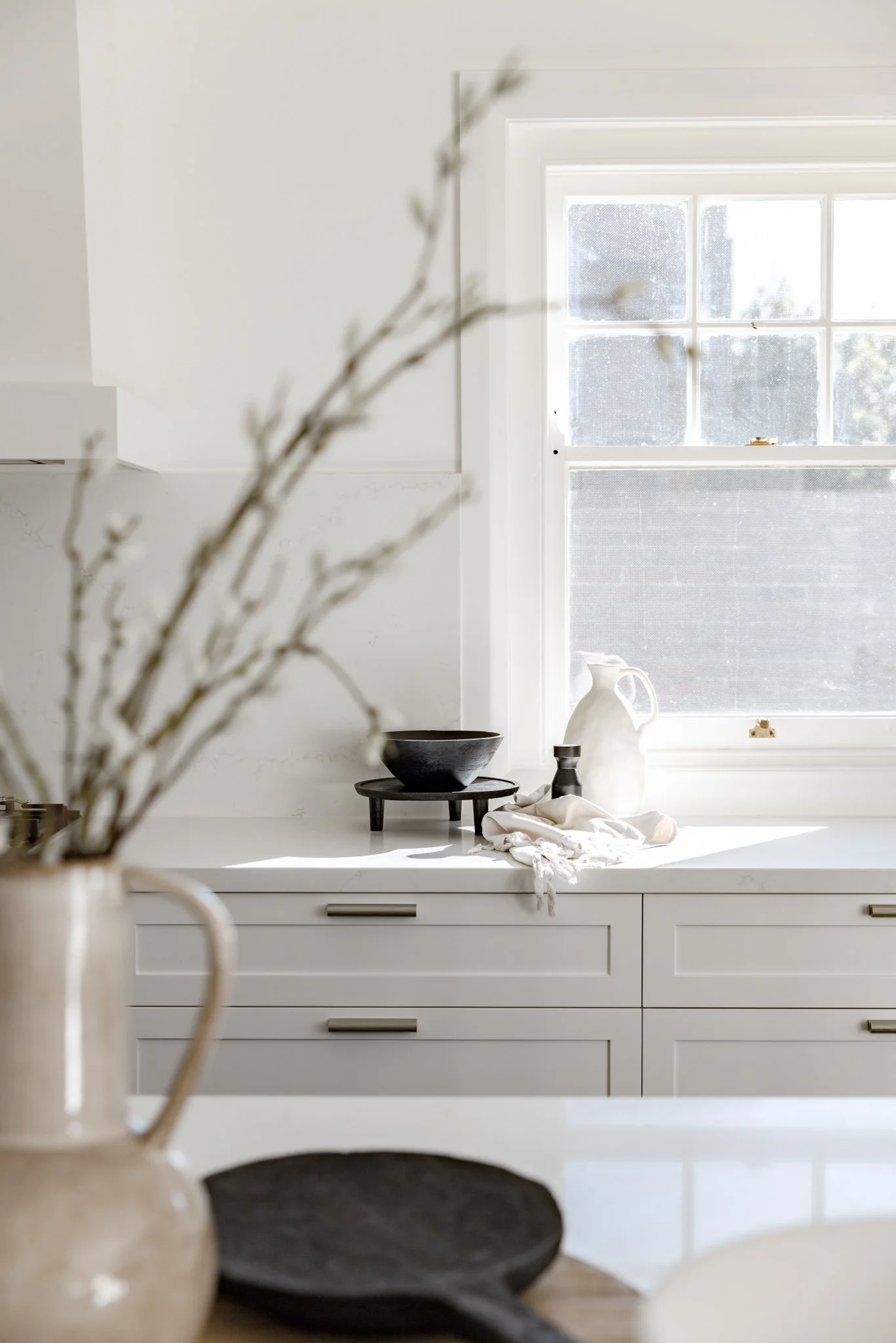Bright kitchen with a white countertop and cabinetry, window with sunlight streaming in, black and white ceramics, and a blurred vase with branches in the foreground.