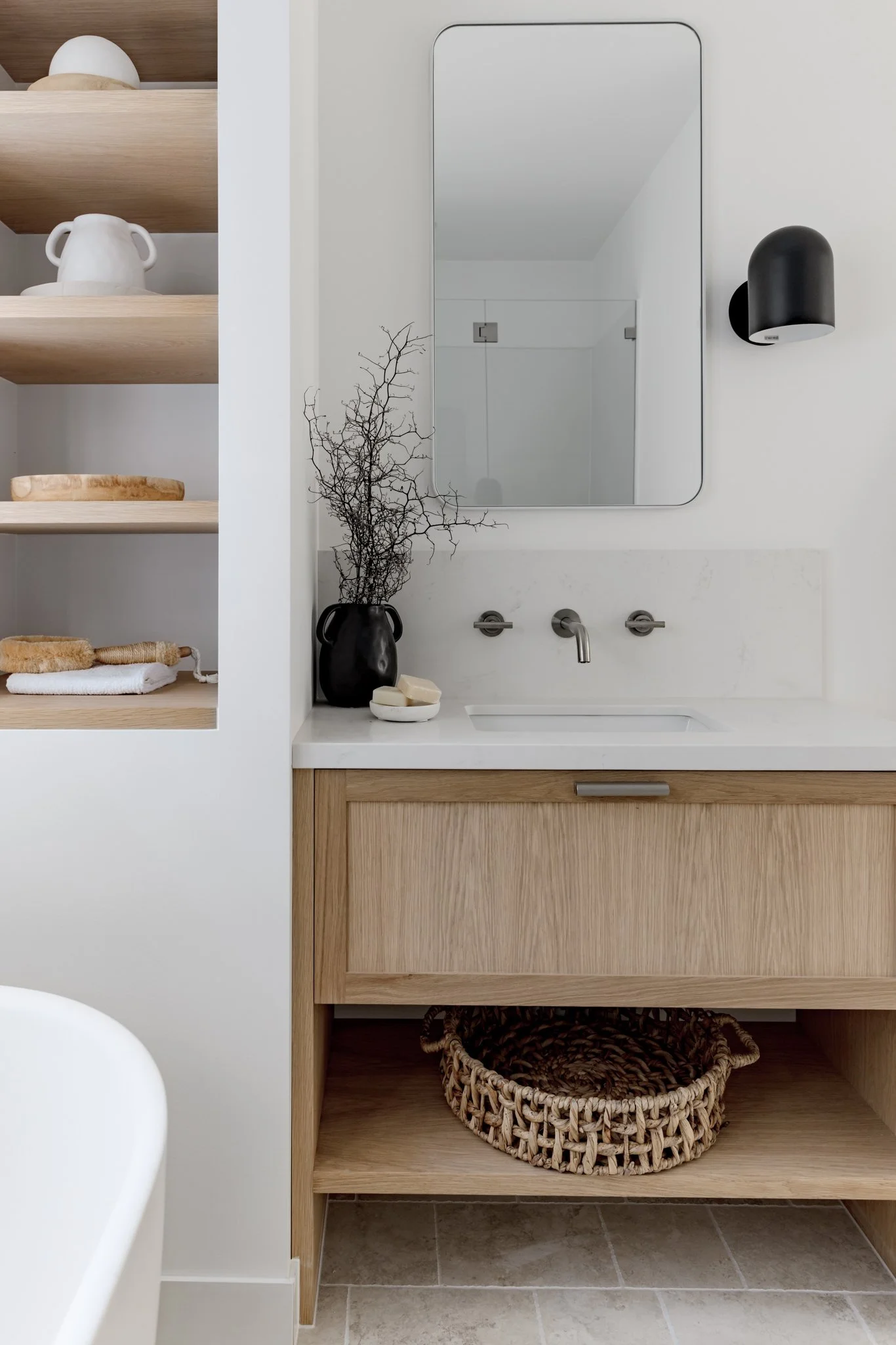 Modern bathroom vanity with a white countertop, wooden cabinet, and woven basket underneath. A black vase with dried branches and a soap dish with soap bars are on the counter. A mirror and a black wall-mounted light fixture are above the sink, and a partially visible bathtub is to the side.