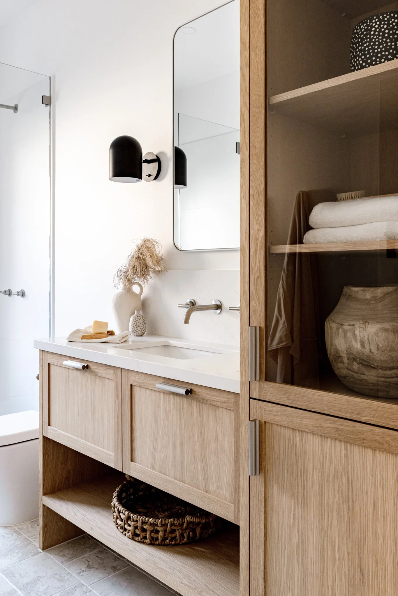 Modern bathroom with wooden vanity, white countertop, and black wall sconces, mirror, and textured decor with natural tones.