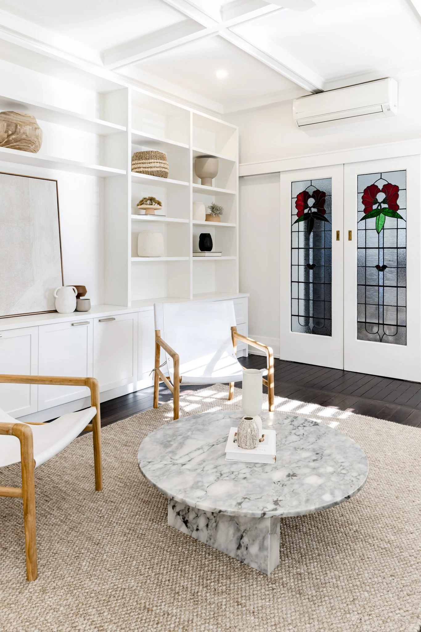 Bright living room with white built-in shelving, stained glass door with floral design, marble coffee table, and white armchairs with wooden frames.
