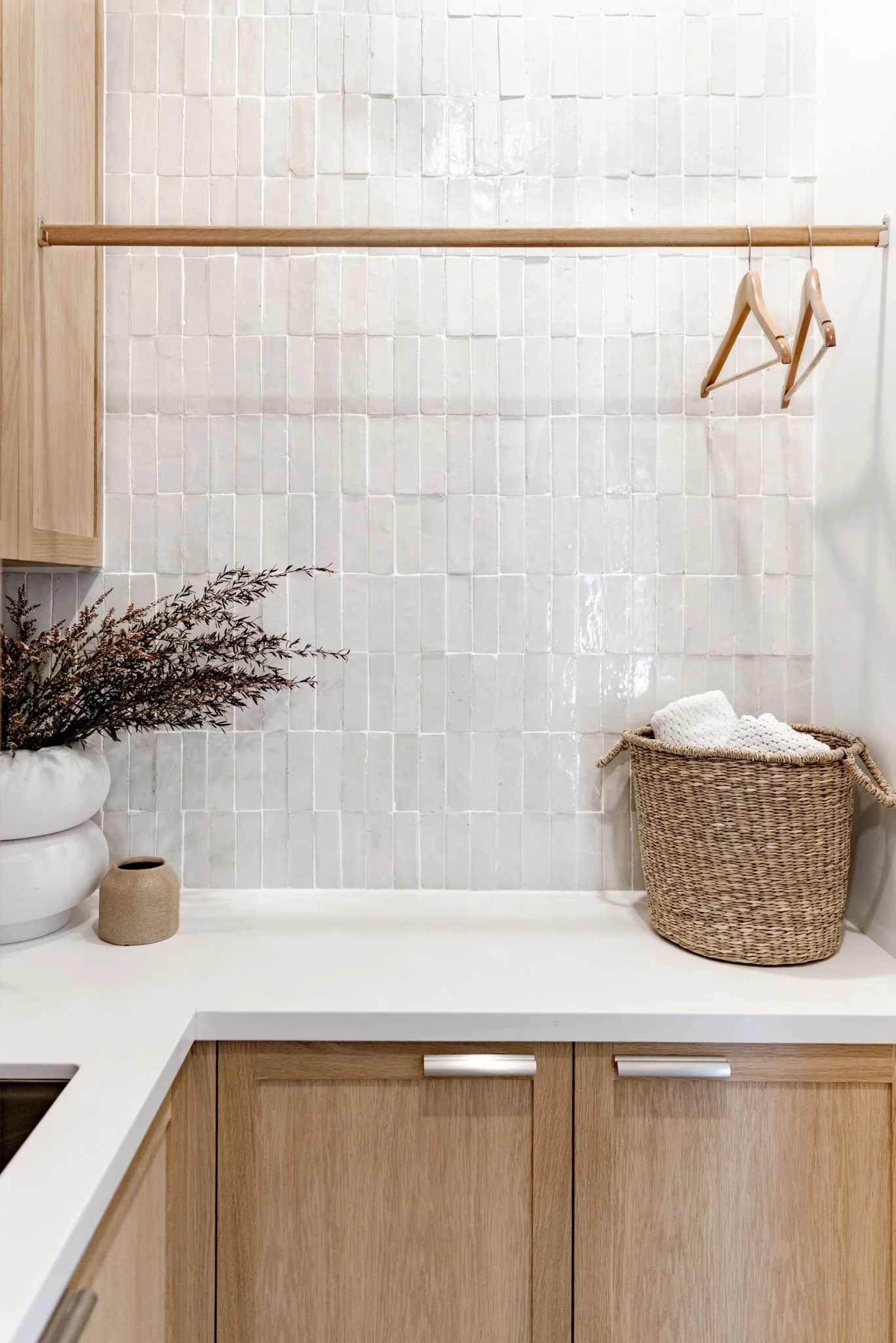 Minimalist kitchen with white tiled backsplash, natural wood cabinets, a white countertop, and woven basket with cloths, decorative vases, and a plant.