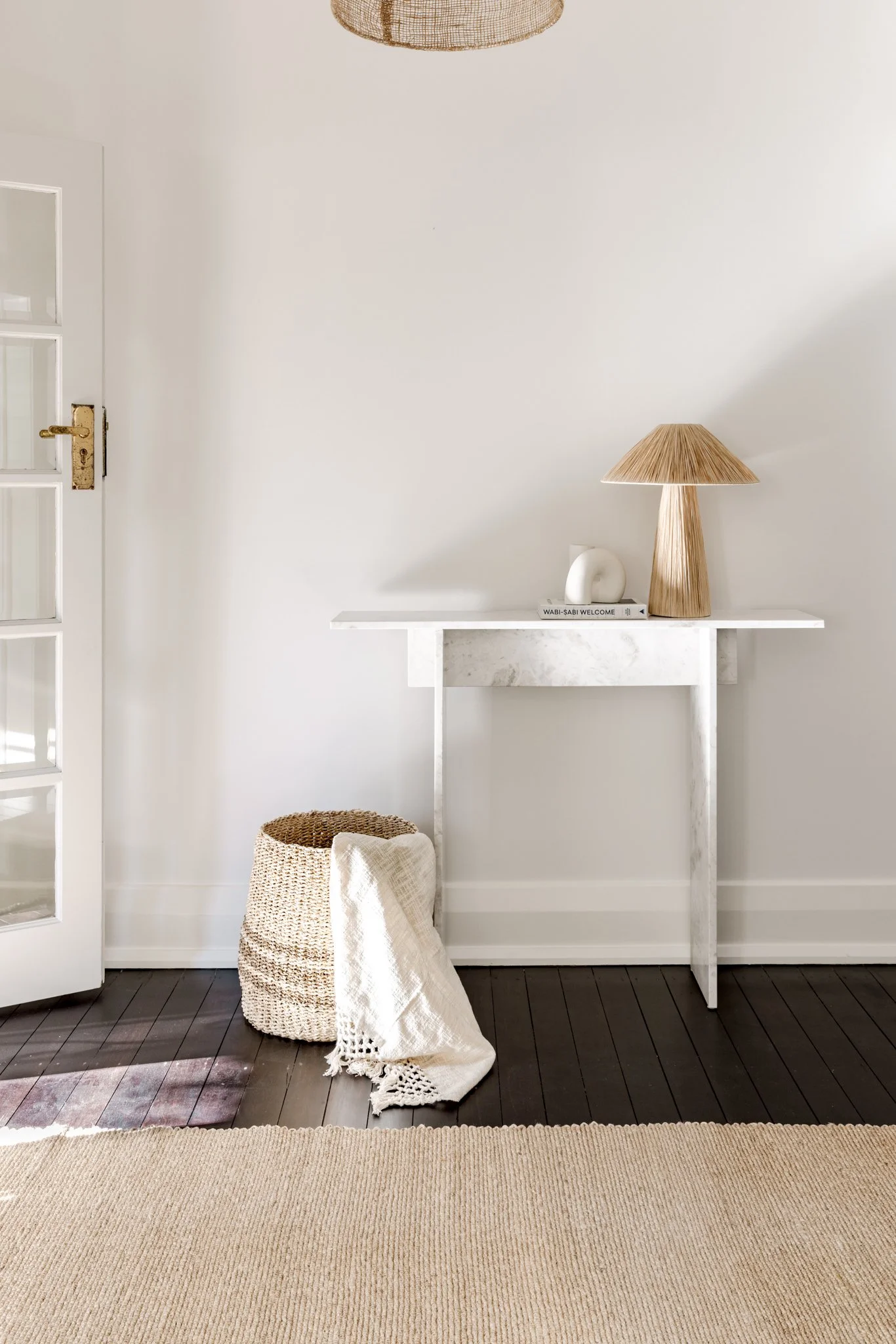 A minimalist entryway with a white marble console table, a woven basket with a white blanket, a beige table lamp, and a white decorative object on a book, all set against a white wall and dark wooden floor.
