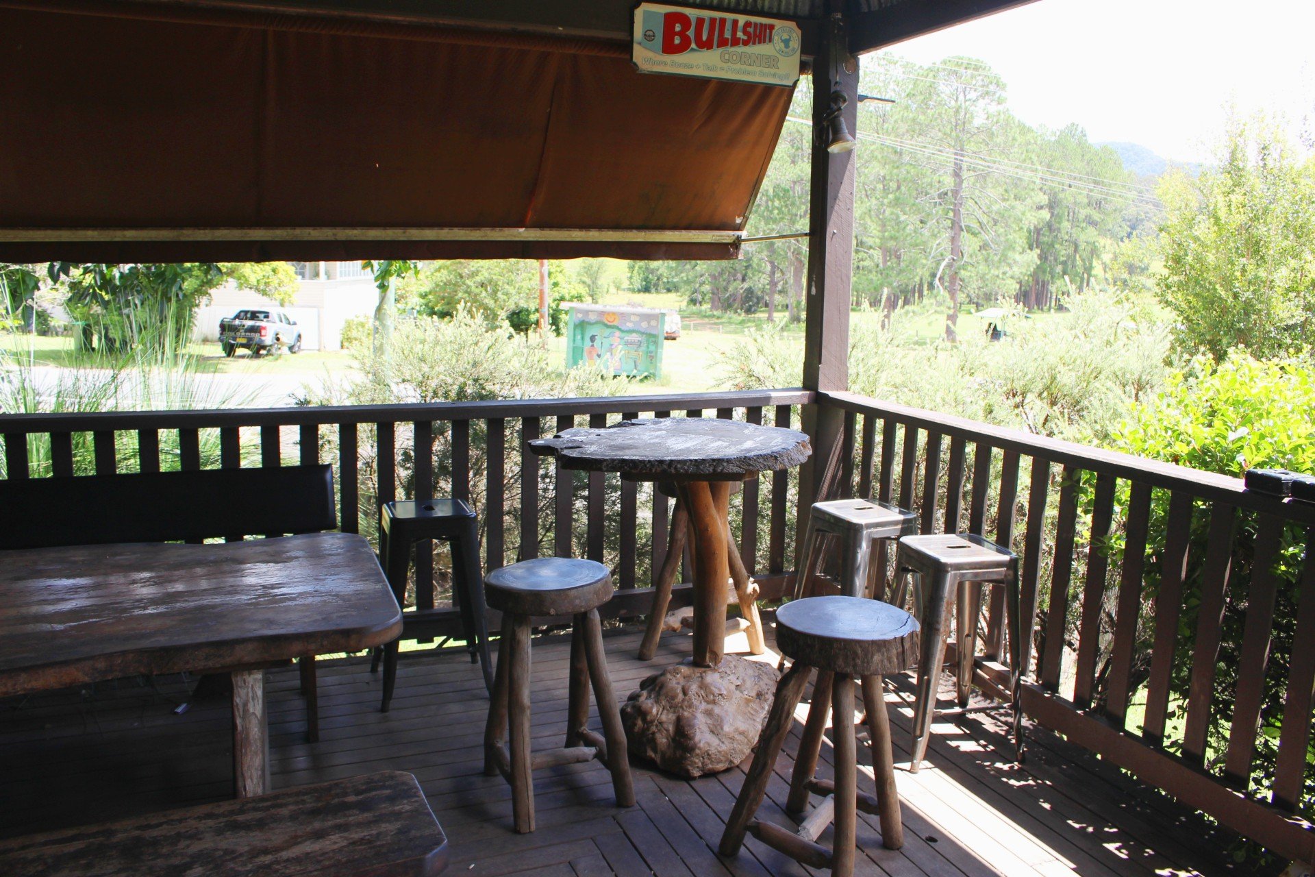 Outdoor seating area on a wooden deck with rustic wooden tables and stools, surrounded by greenery and trees, and a sign that reads 'BULLSHIT CORNER' hanging overhead.