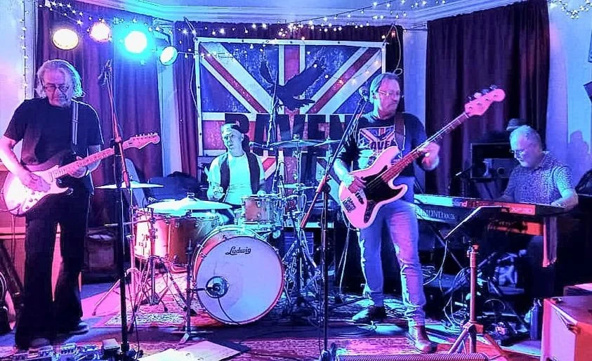 Four musicians perform on a stage with a British-themed backdrop, including a man playing guitar, a drummer, a man on bass guitar, and a woman on keyboard. The scene is illuminated with colorful stage lights.
