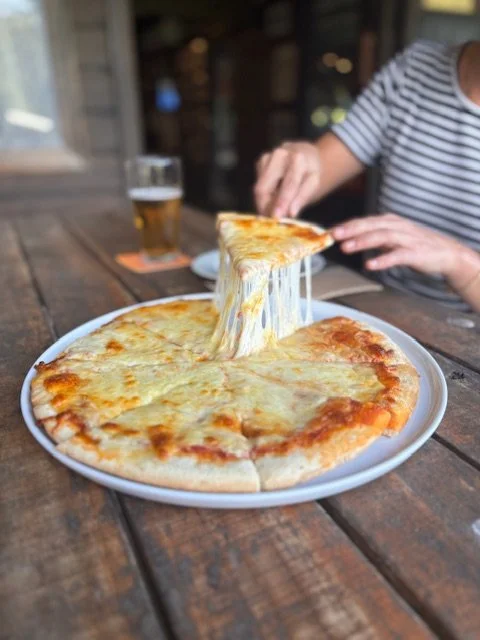 A cheese pizza on a white plate on a wooden table, with a person in a striped shirt lifting a slice and a glass of beer in the background.