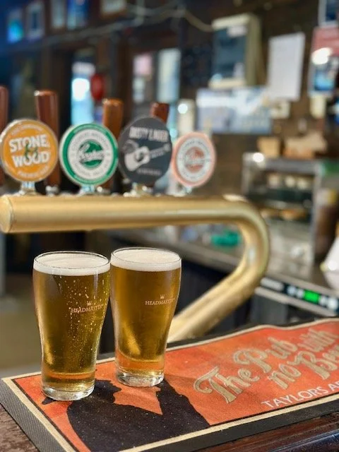Two glasses of beer on a bar counter with beer taps in the background.