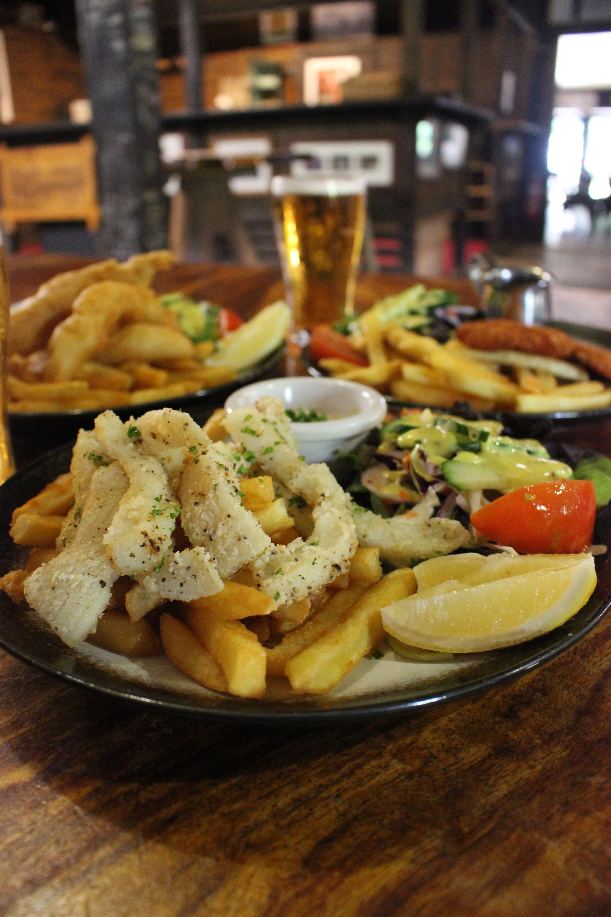 A plate of fish and chips with lemon wedges, a side salad, and tartar sauce, with a glass of beer in the background, on a wooden table in a restaurant setting.