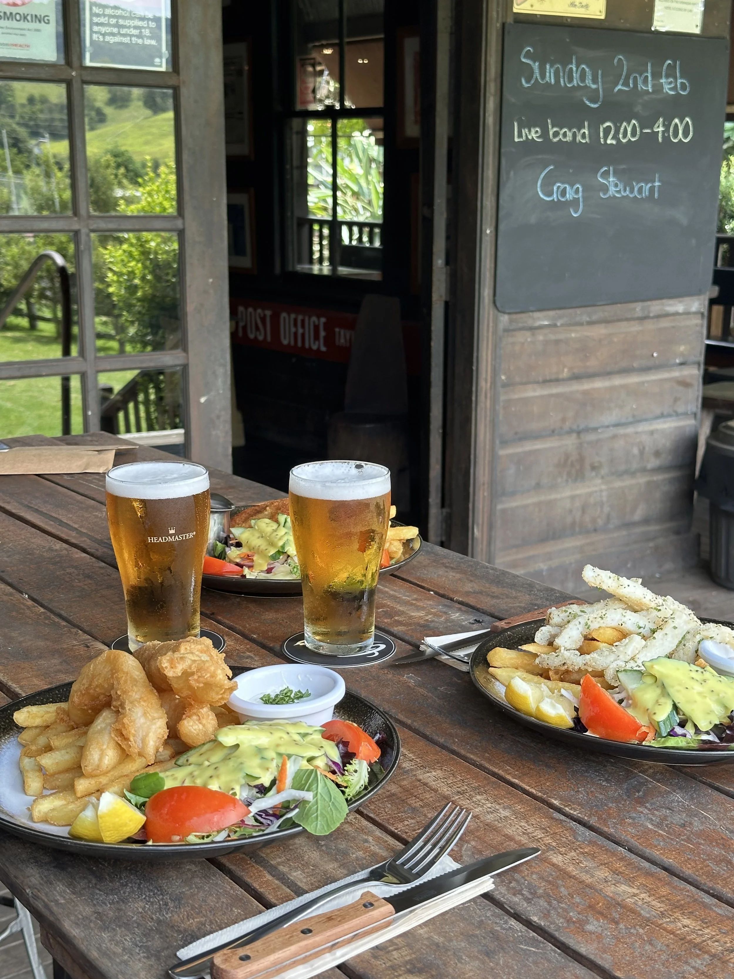 Outdoor table with two plates of fish and chips, salad, and two glasses of beer, set near a rustic wooden building with a chalkboard sign and a window showing greenery outside.